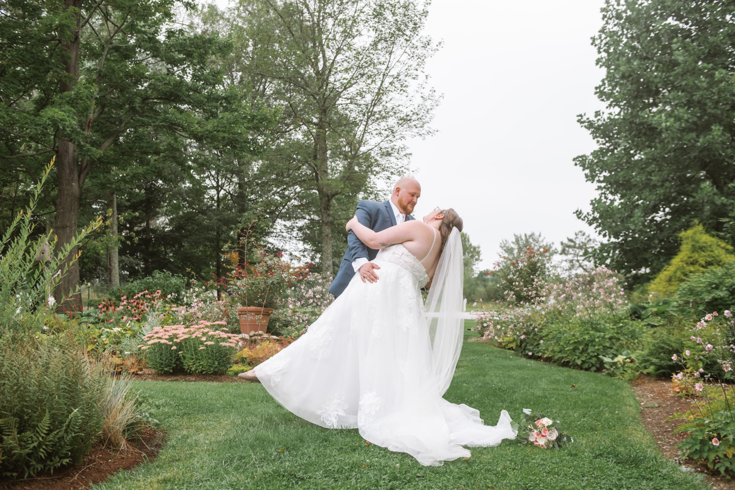 Groom dipping his bride romantically in a garden. Her bouquet is set to the behind her dress train and veil on the ground.  She is wearing a long white lace gown with a long veil and he is wearing a blue suit with a white tie. 