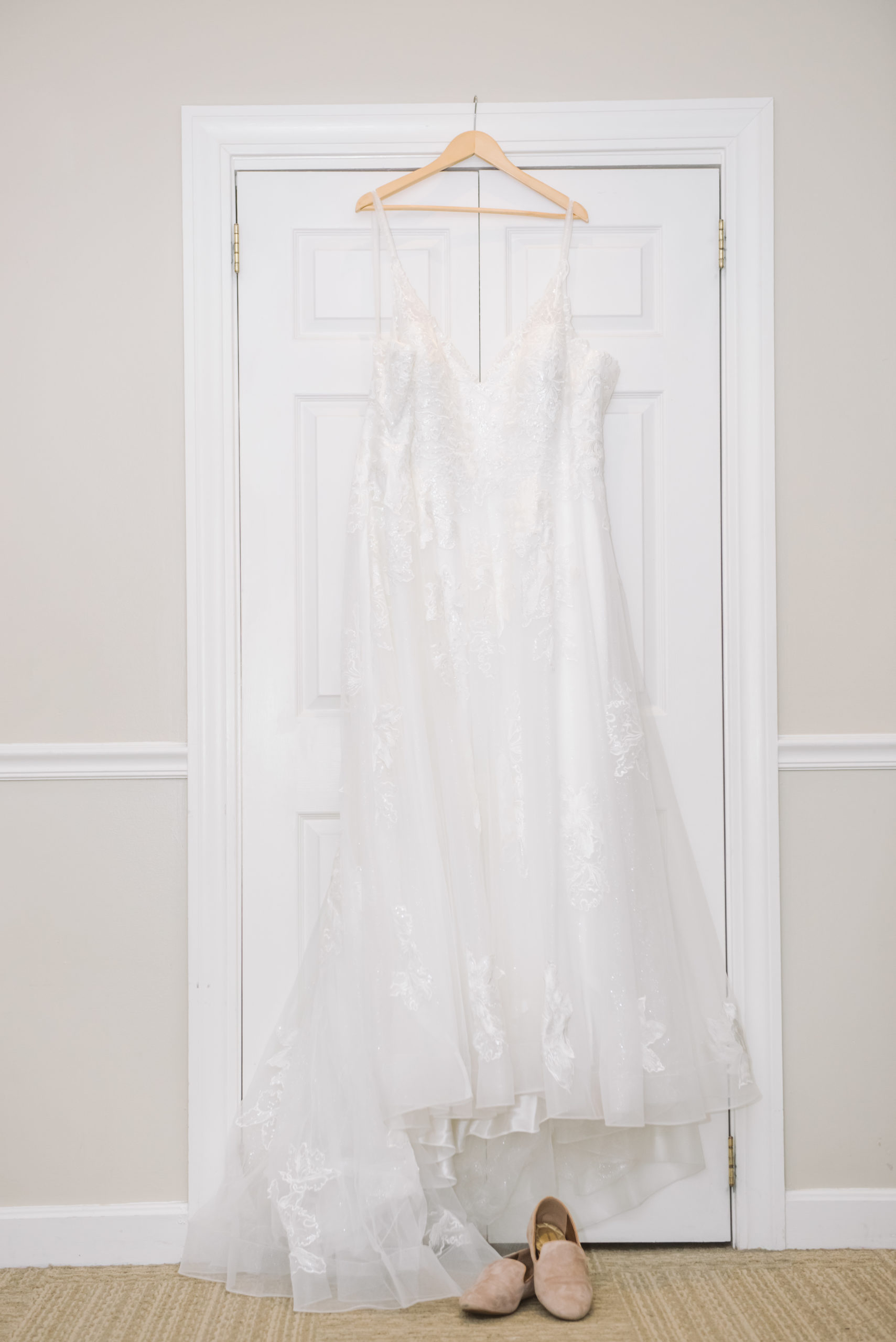 Bride's ceremony gown hanging on a light wooden hanger on a white closet door. Her ceremony shoes are set below the dress. They are a muted pink/beige velvet.