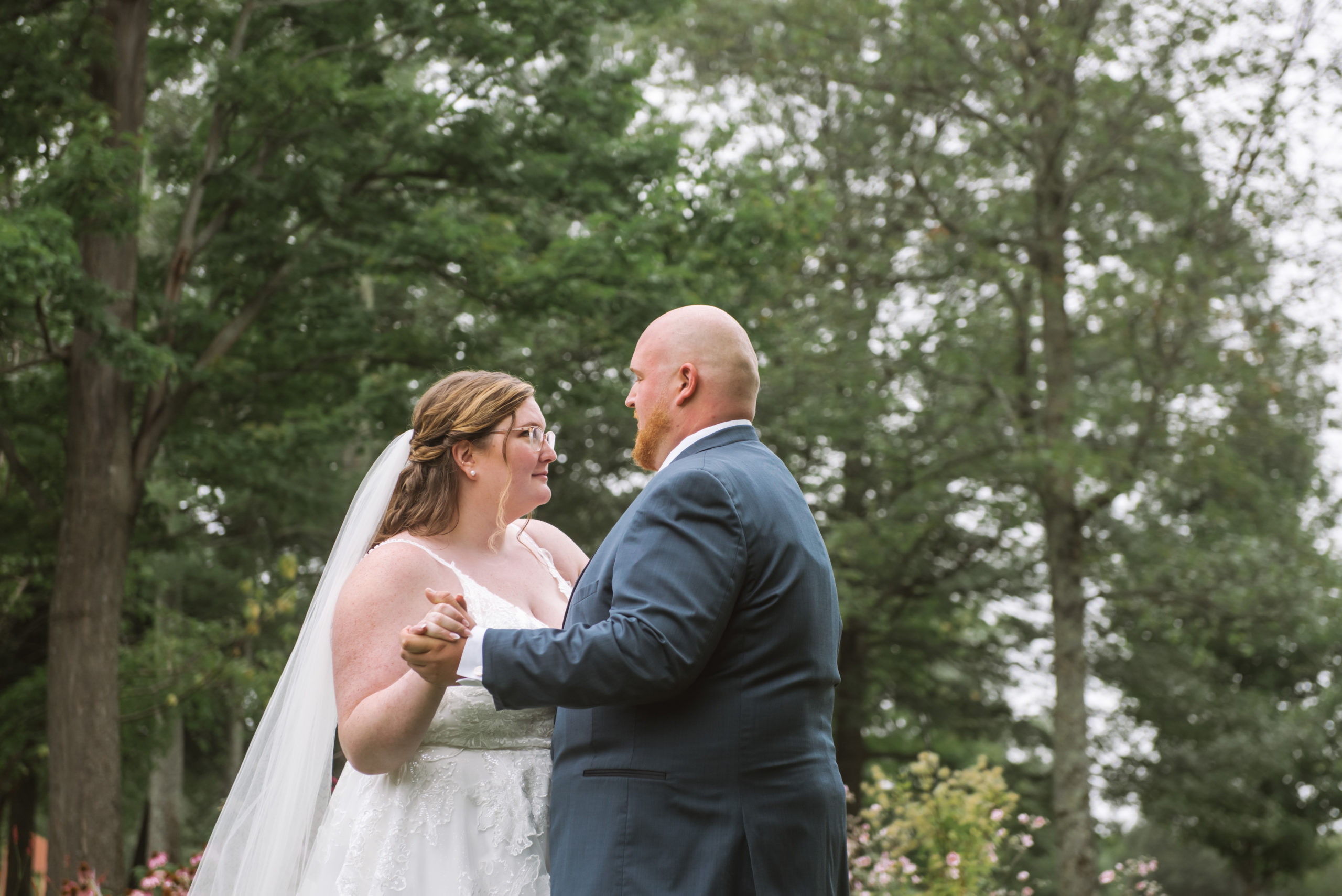 Bride and groom standing facing one another with their arms wrapped around each other and their hands in each other's hands in a slow dance. They are looking into each other's eyes. There are trees in the background.