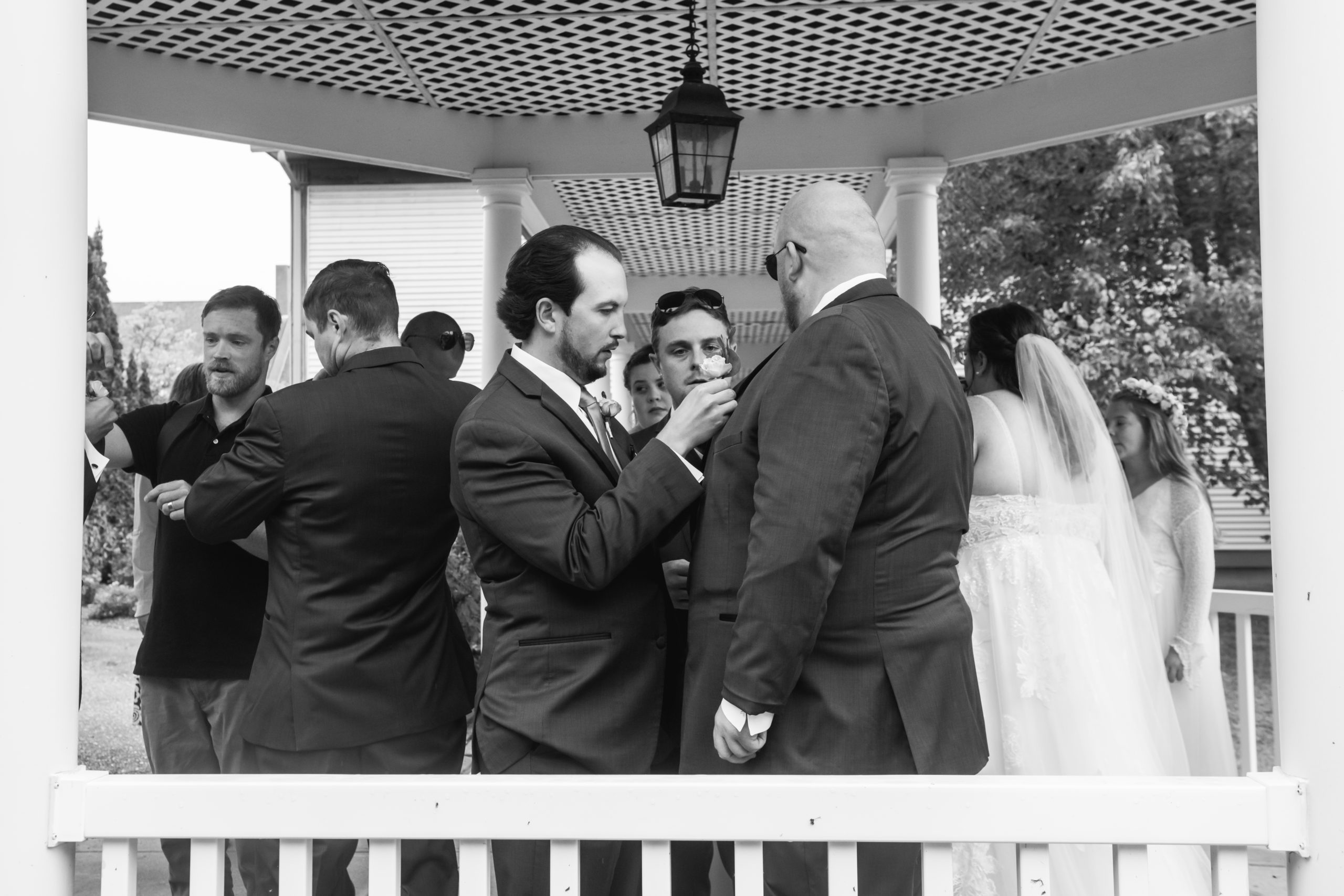 Black and white photo of one of the groomsmen helping the groom put on his boutonniere under a covered walkway. There are other members of the wedding party all around.