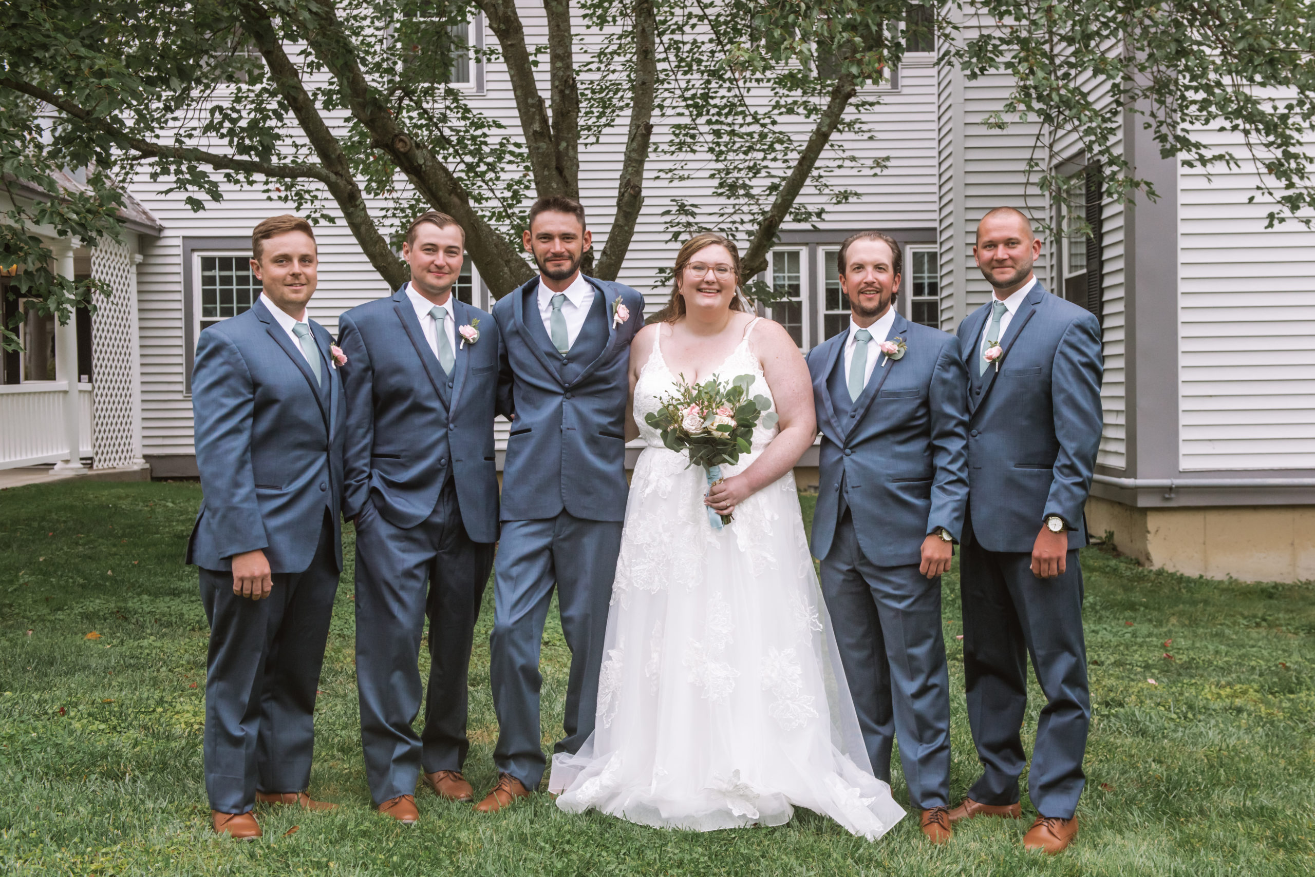 Bride standing in line with all of the groomsmen. She is wearing a long white lace gown with a long veil and is holding her bouquet in her left hand. The groomsmen are all wearing blue suits with light green ties and boutonnieres.