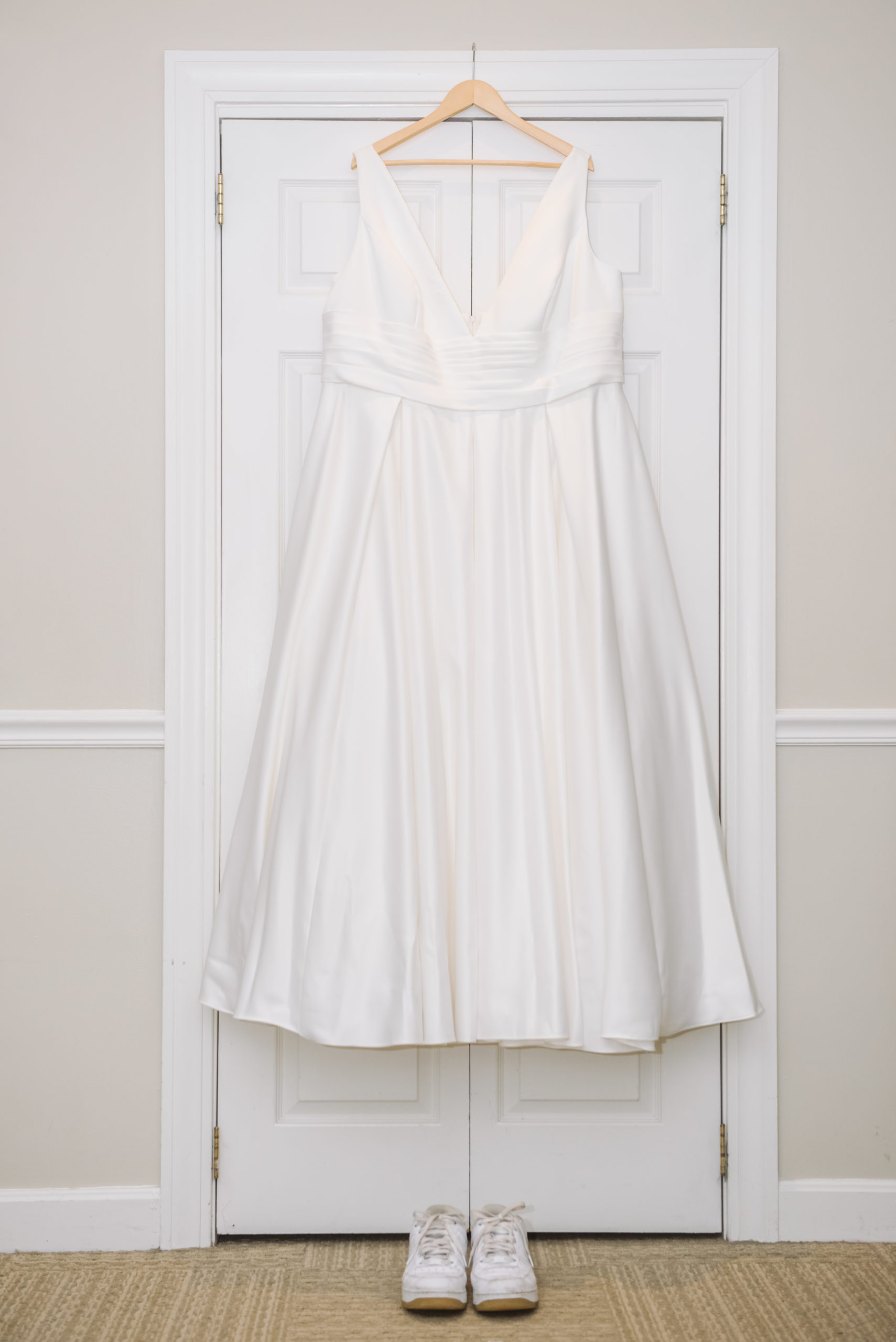 Bride's reception gown hanging on a light wooden hanger on a white closet door. It is a deep v-neck full-length gown with no train. Her reception shoes are set below the dress. They are air force one white sneakers.