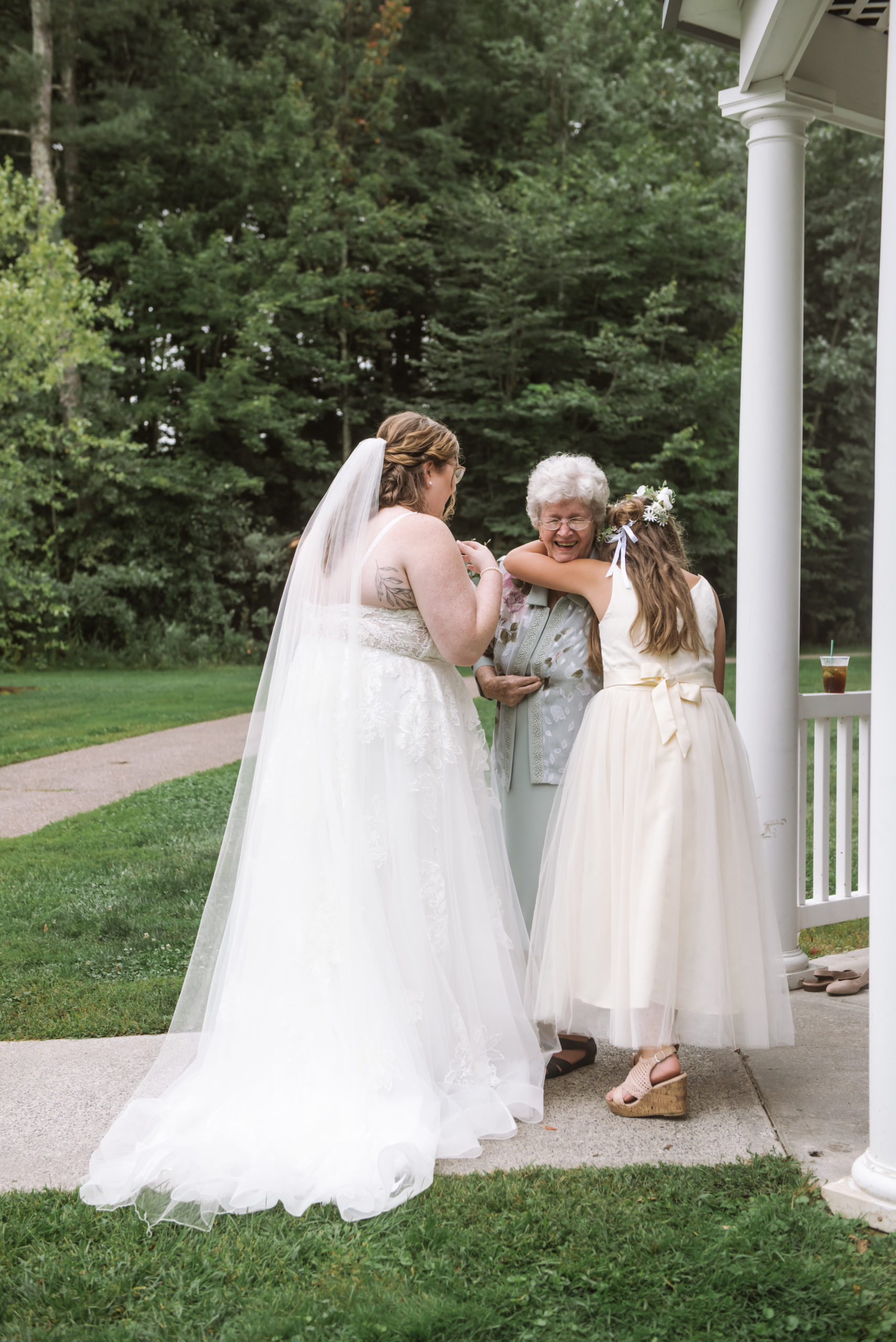 Tender moment with the bride, her grandmother, and one of the bride's younger family members. The younger family member is actively hugging the bride's grandmother who is smiling open-mouthed. The bride is facing toward her grandmother, away from the camera. There are trees in the background.