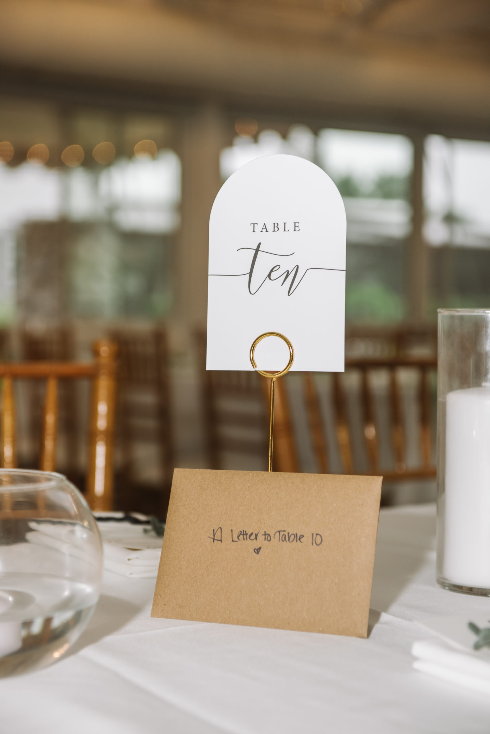 Close up of the "TABLE ten" placecard and a brown envelope with the note "A Letter to Table 10 [illustrated heart]" written on the front. They are set on a white tablecloth. There is an unlit candle behind and to the left. Reception chairs are visible in the background.