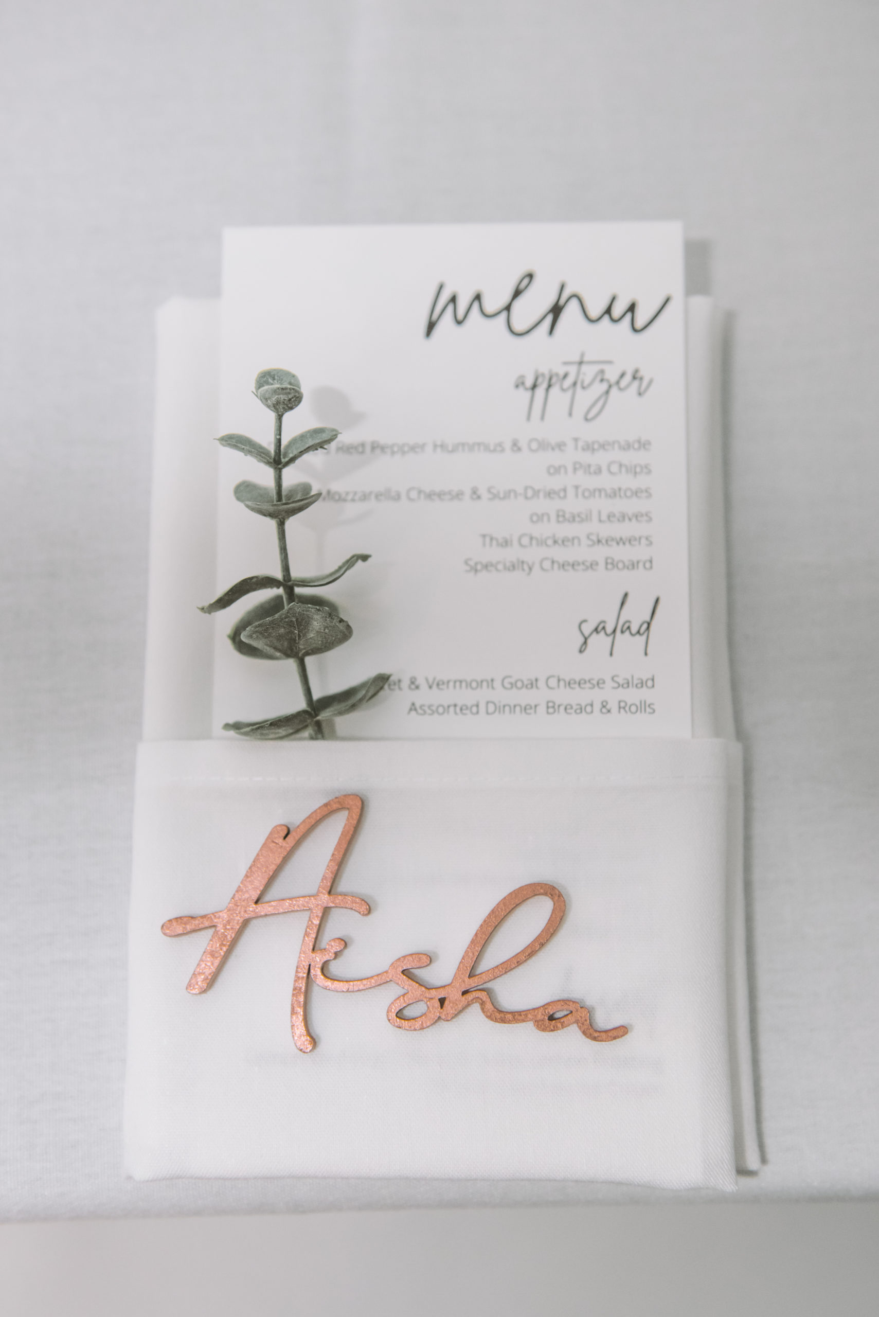 Detail of an individual placesetting for "Aisha" written out as a wooden namecard atop a white cloth napkin which has the menu and a piece of greenery in its pocket.