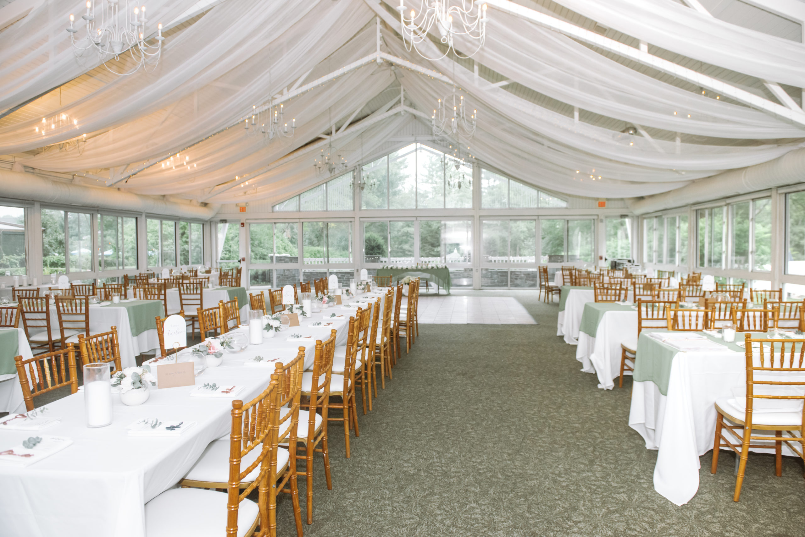Reception venue full of set tables, hanging white sheer curtains on the ceiling, small chandeliers hanging from the ceiling, and a dance floor in the far section of the room.