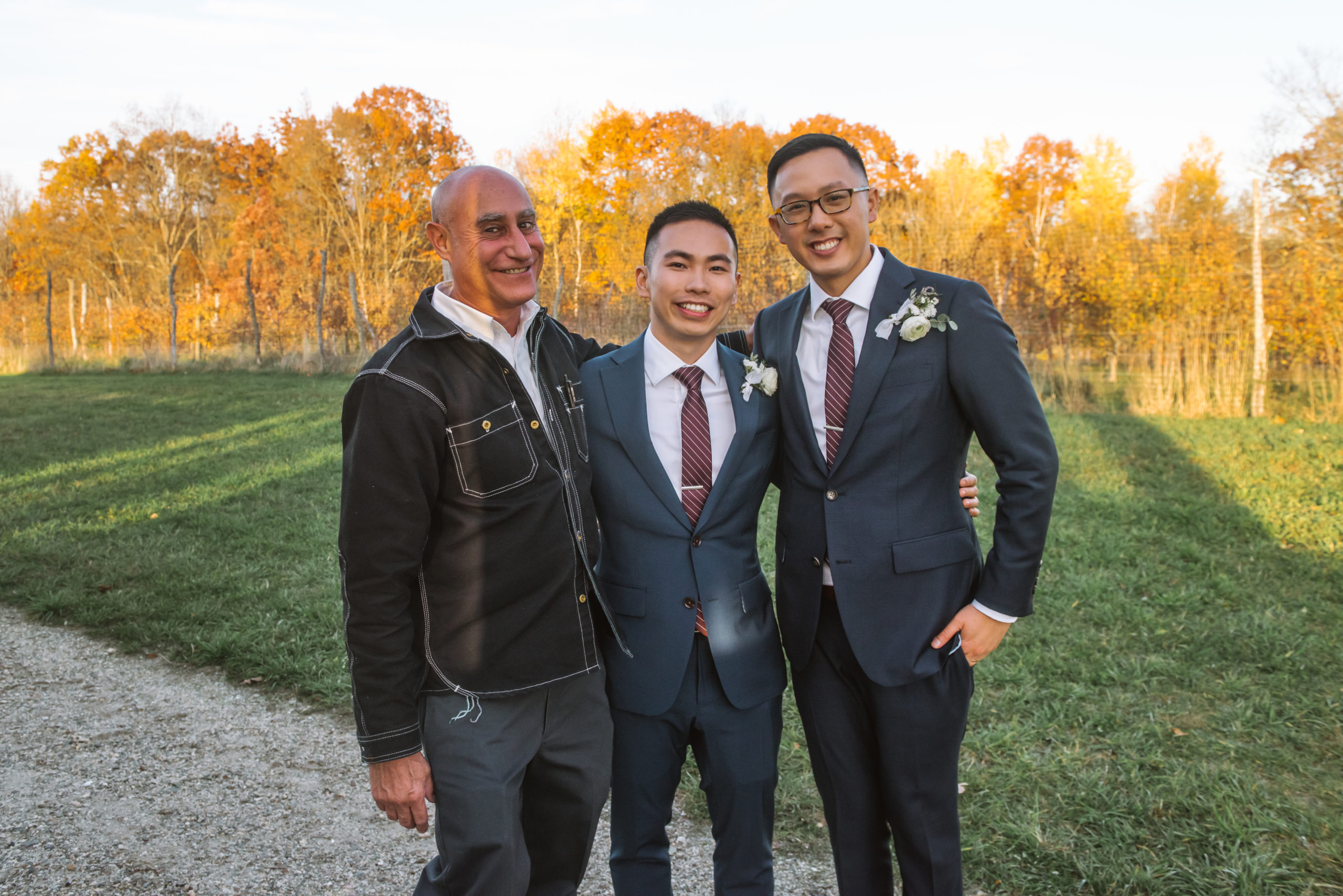 Two grooms standing side by side with their wedding planner. They all have their arms around one another and are smiling directly to the camera. There are autumnal trees in the background.