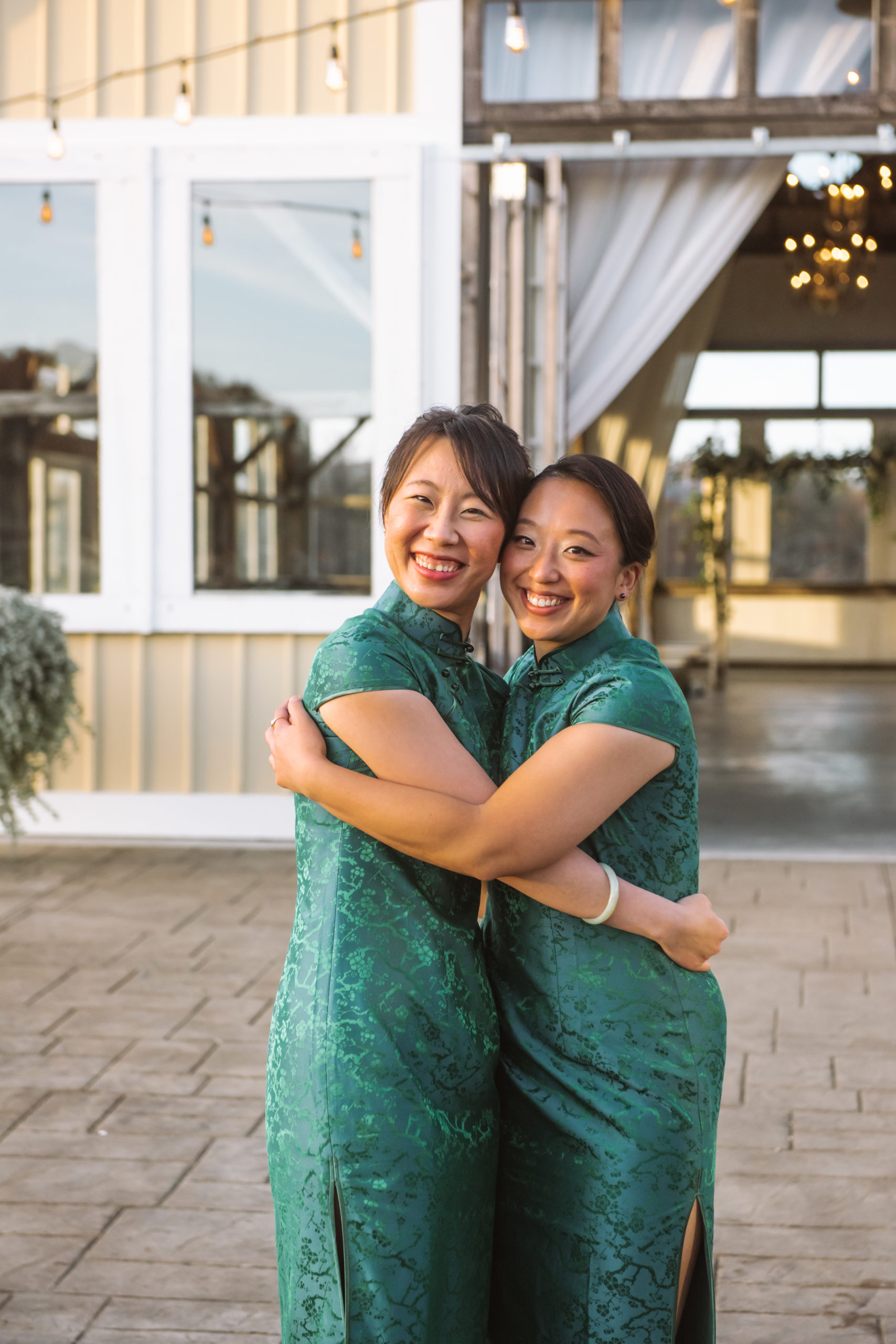 Two women who are part of the wedding party, dressed in dark green qi paos, facing one another with their heads turned to look directly at the camera. Their arms are wrapped around one another in a hug and they are both smiling. They are standing in the stone courtyard area in front of the ceremony location.