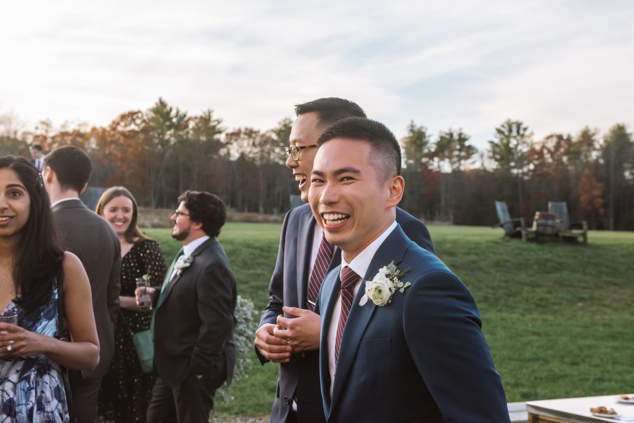 Groom's head is turned toward the camera and he is smiling/laughing. His husband is also smiling/laughing behind him, looking straight ahead (he is faced towards his guests). He has his right hand holding his wedding band on his left hand.