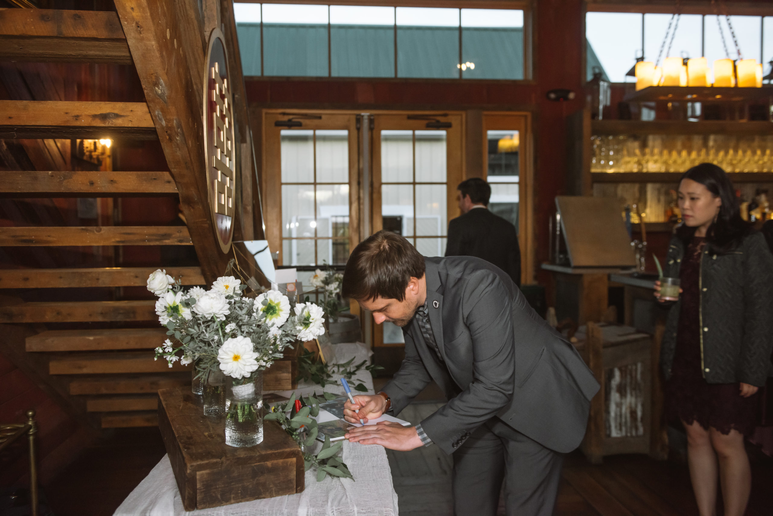 Wedding guest mid-signing of the guest book. There is a bouquet of white flowers in a vase on top of a wooden platform on the table.