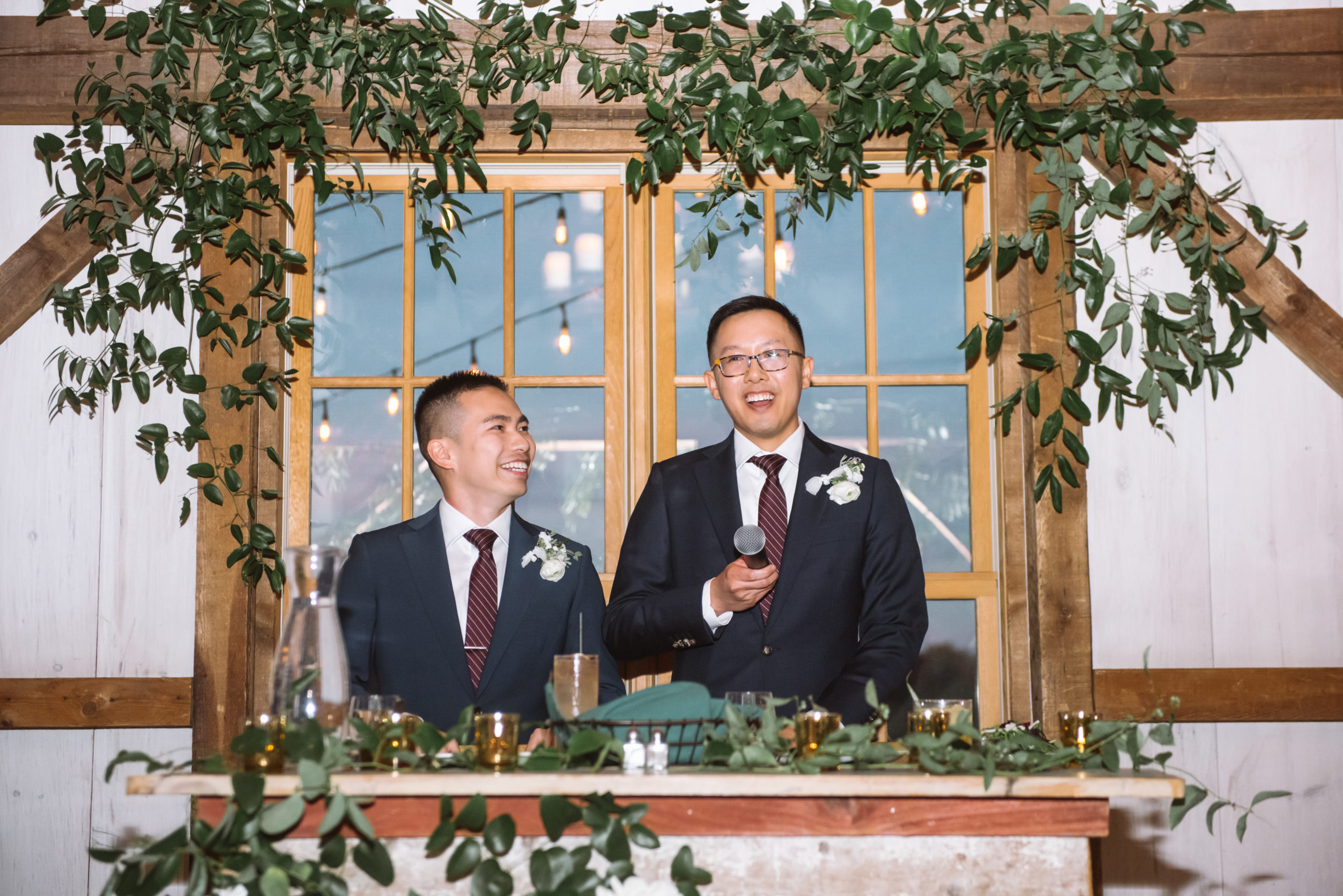 Two grooms standing about to give a speech to begin their reception. One groom is looking off to his right, mouth open in a smile. He is holding a microphone. His husband is standing to his right and is looking up towards him with a big smile on his face. They are standing at their couple head table which is decorated with greenery as well as the wooden beam above them.