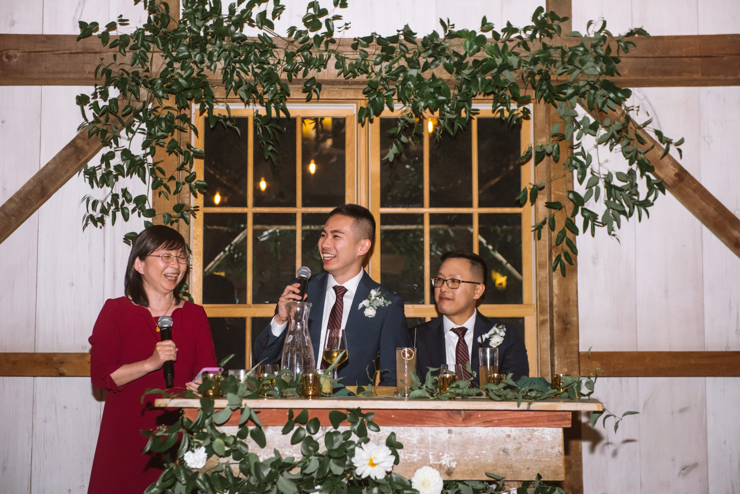 Two grooms at the head couple table (which is decorated with greenery as well as the wooden beam above them) with one of the groom's mothers. One of the grooms and his mother are giving a speech. One groom is looking off to his right, mouth open in a smile. He is holding a microphone. His husband is sitting to his left and is looking towards him with a soft smile.