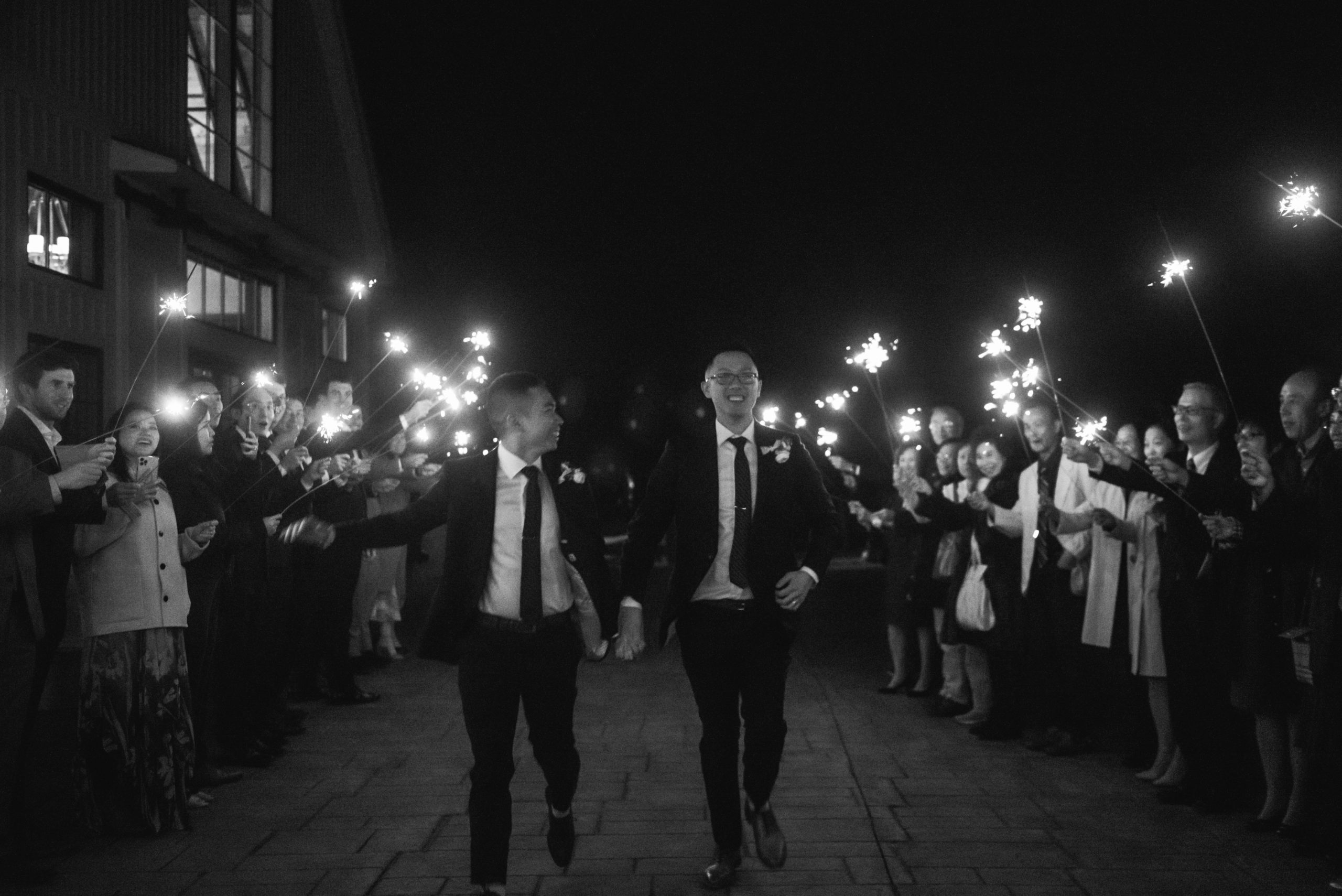 Black and white photo of two grooms running hand in hand through an aisle of sparklers held up by their guests.