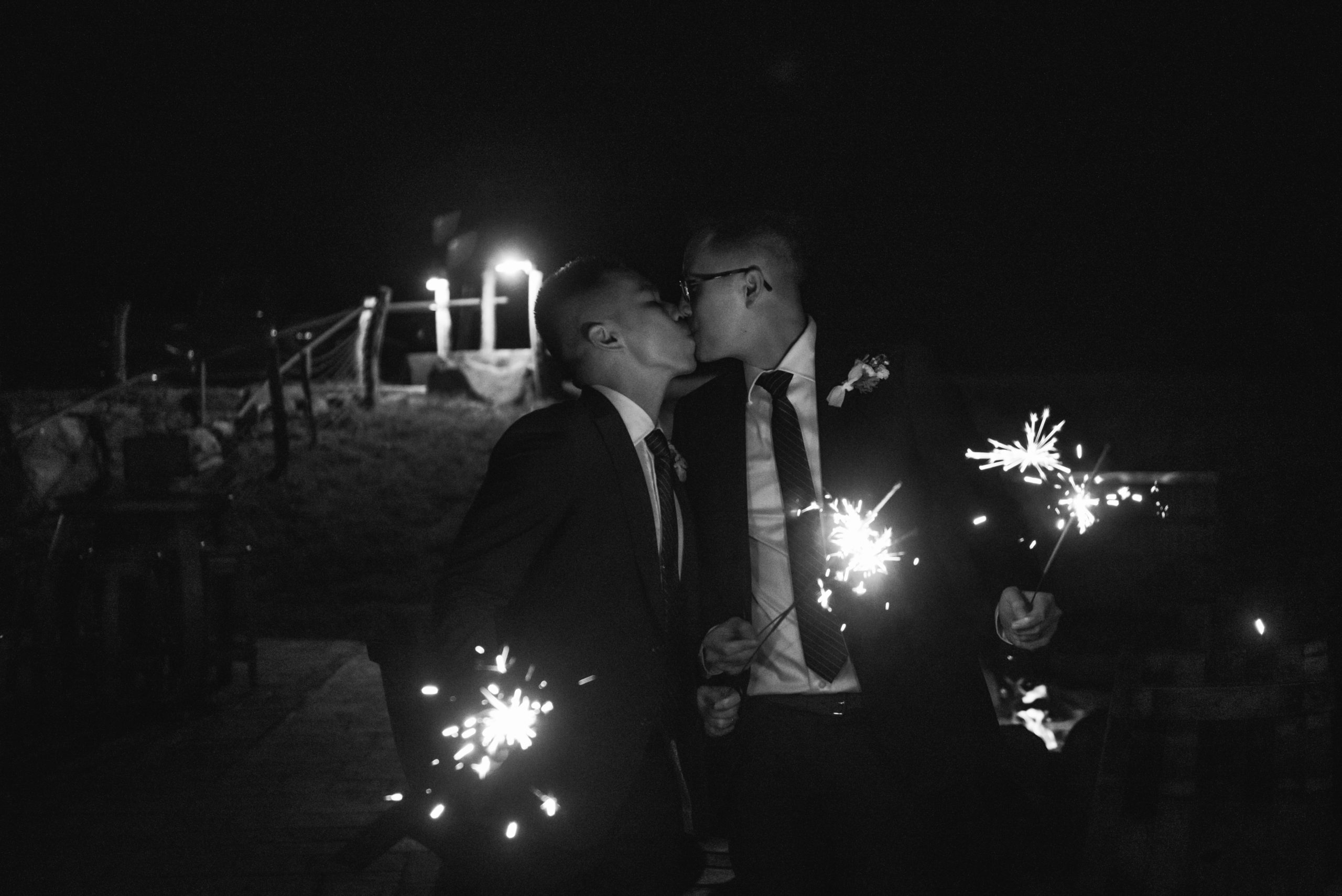Black and white photo of two grooms standing, kissing one another while each holding two sparklers.