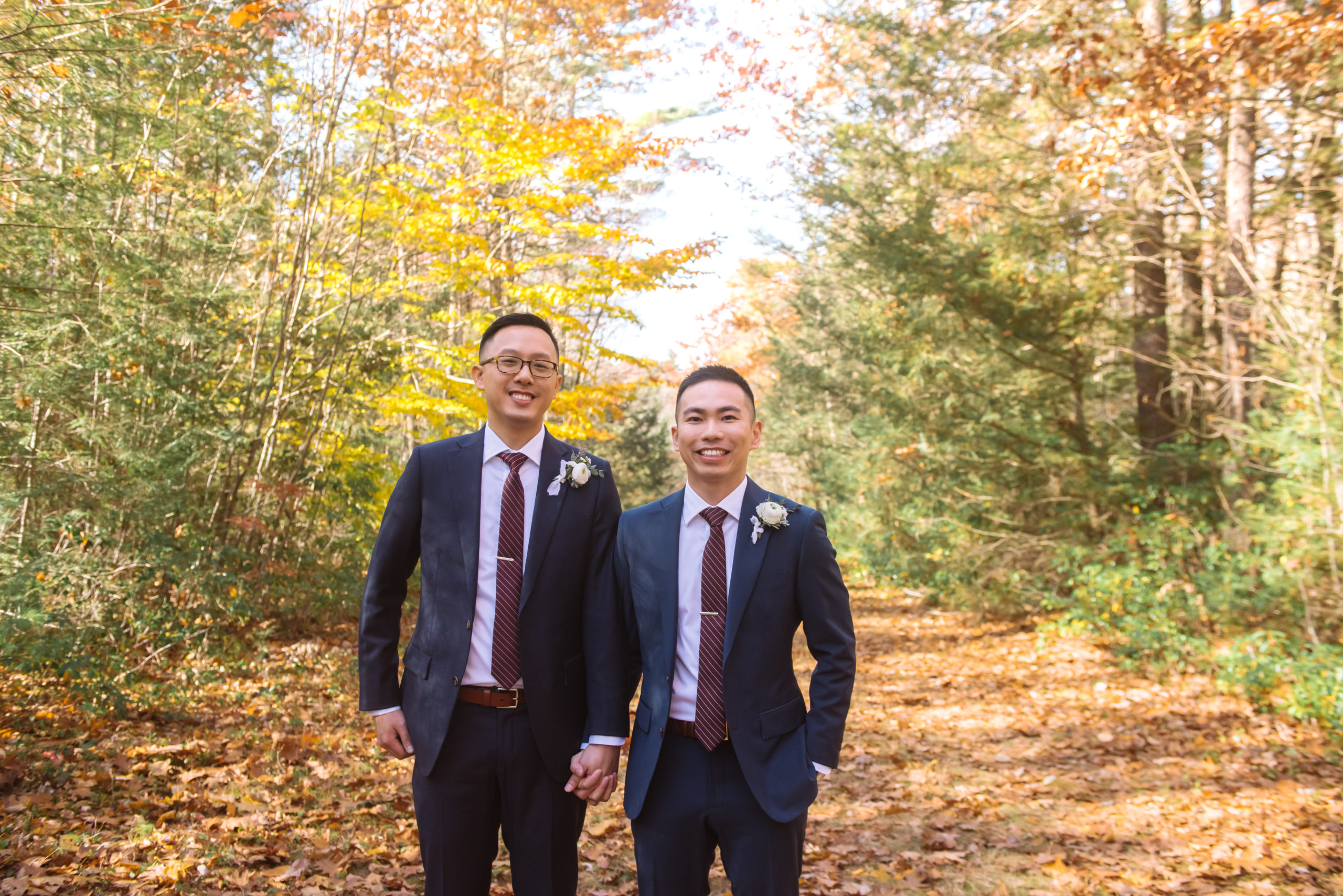 Two grooms standing side by side, holding hands. They are both looking straight to camera and are smiling.  They are dressed in two different colored dark blue suits, have matching striped burgundy ties, and have similar styles of white floral and greenery boutonnieres. They are on an autumnal-leaf-covered trail and there are trees in the background.