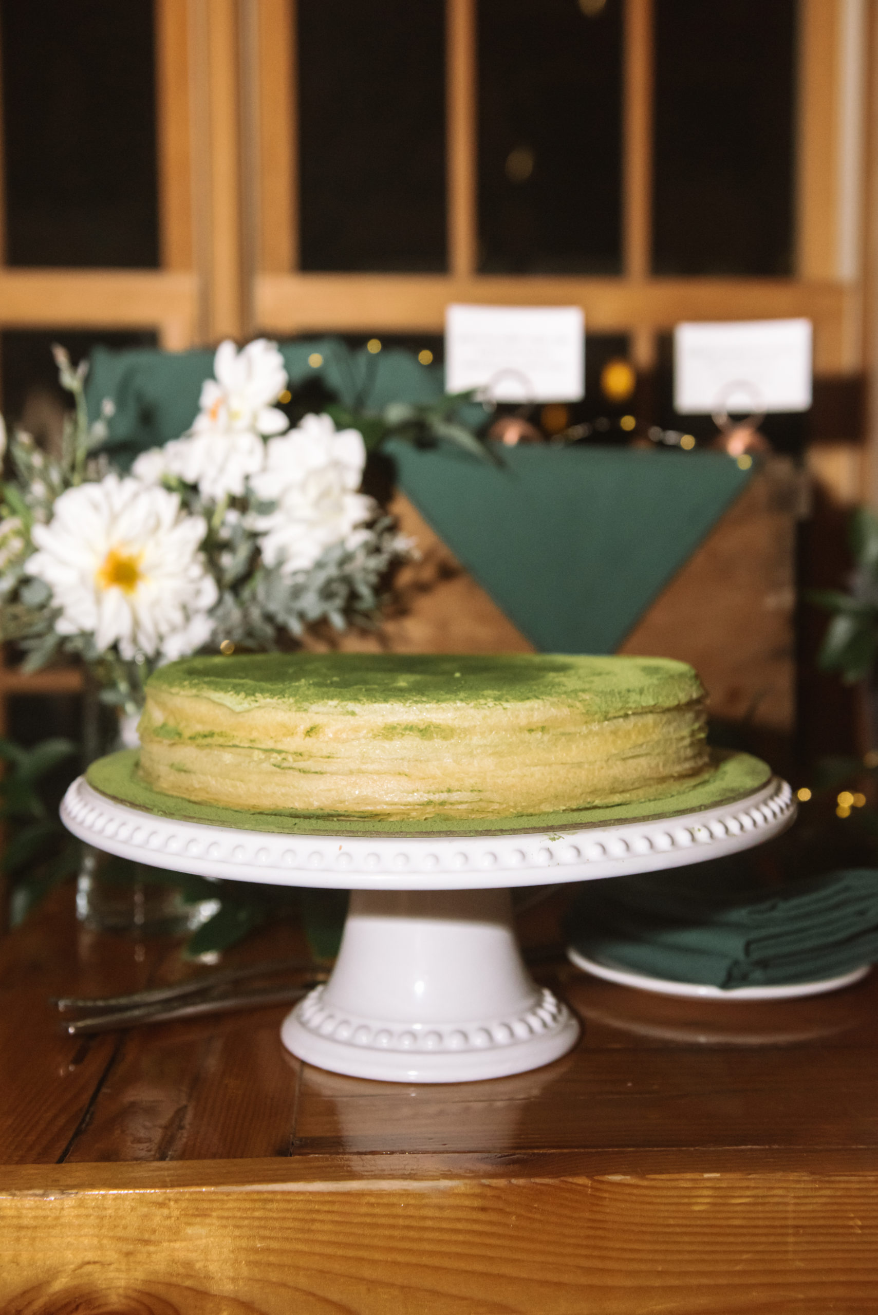 Matcha thousand layer crepe cake on a white cake stand. There is a bouquet of white flowers behind the cake and dark green napkins decorating the wooden table.
