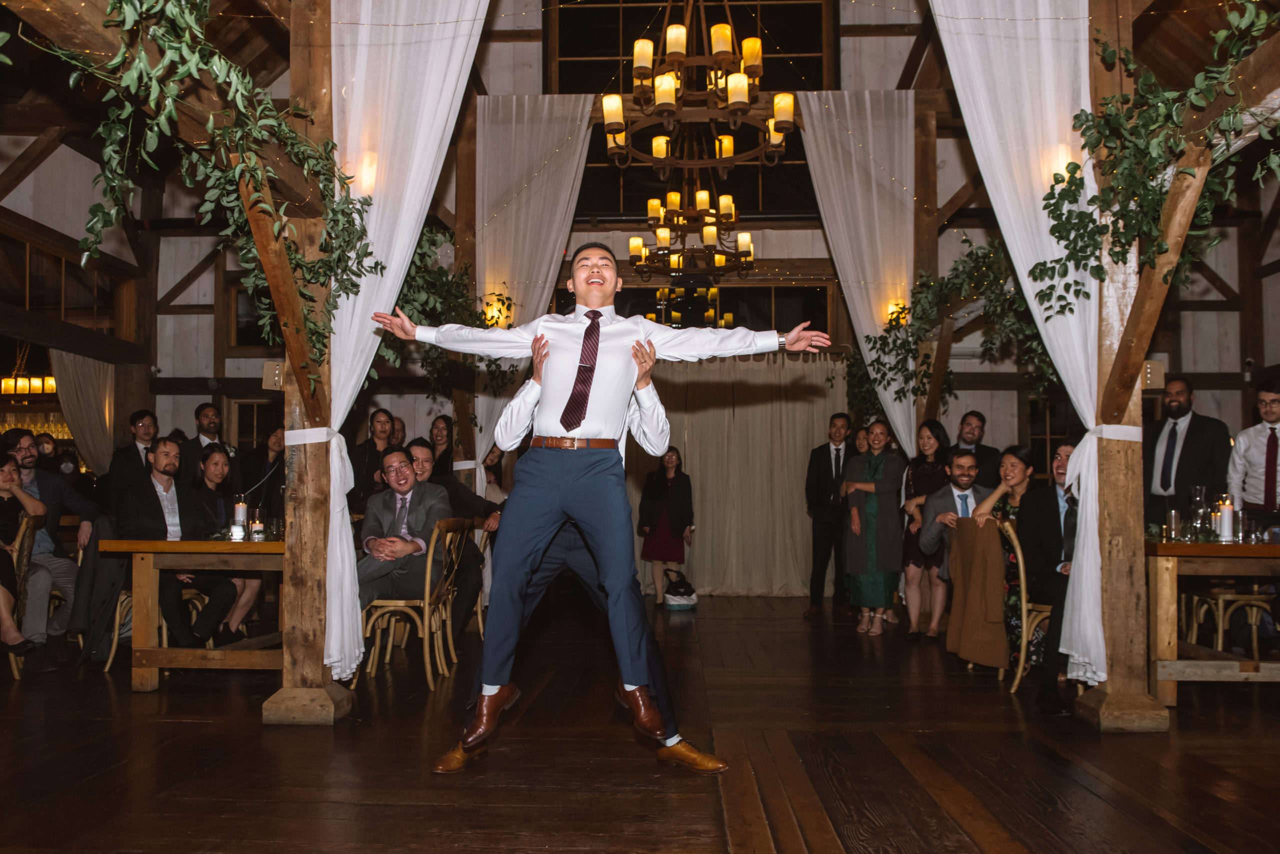 Two grooms having their first dance. One is picking up the other who has his arms outstretched in a wide stance. Their guests are all looking at them and smiling.