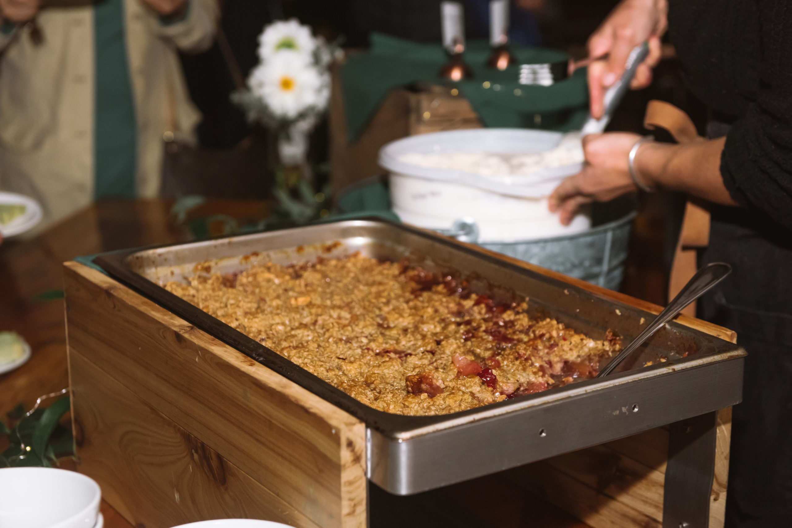 Detail of a tray of apple crumble pie and in the background is someone scooping a ball of vanilla ice cream from the ice cream tub.