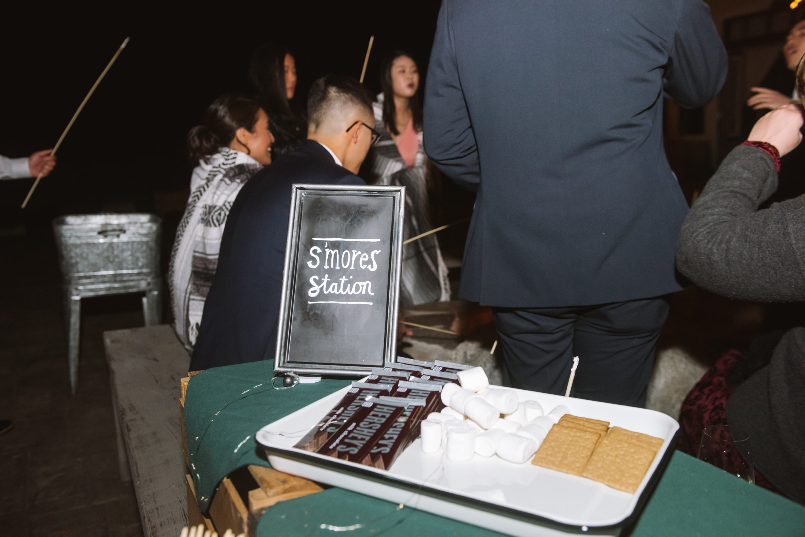 The s'mores station with a blackboard style sign that says "s'mores station" with a tray of Hershey's chocolate bars, marshmallows, and graham crackers. People are behind the station in various stages of eating s'mores.