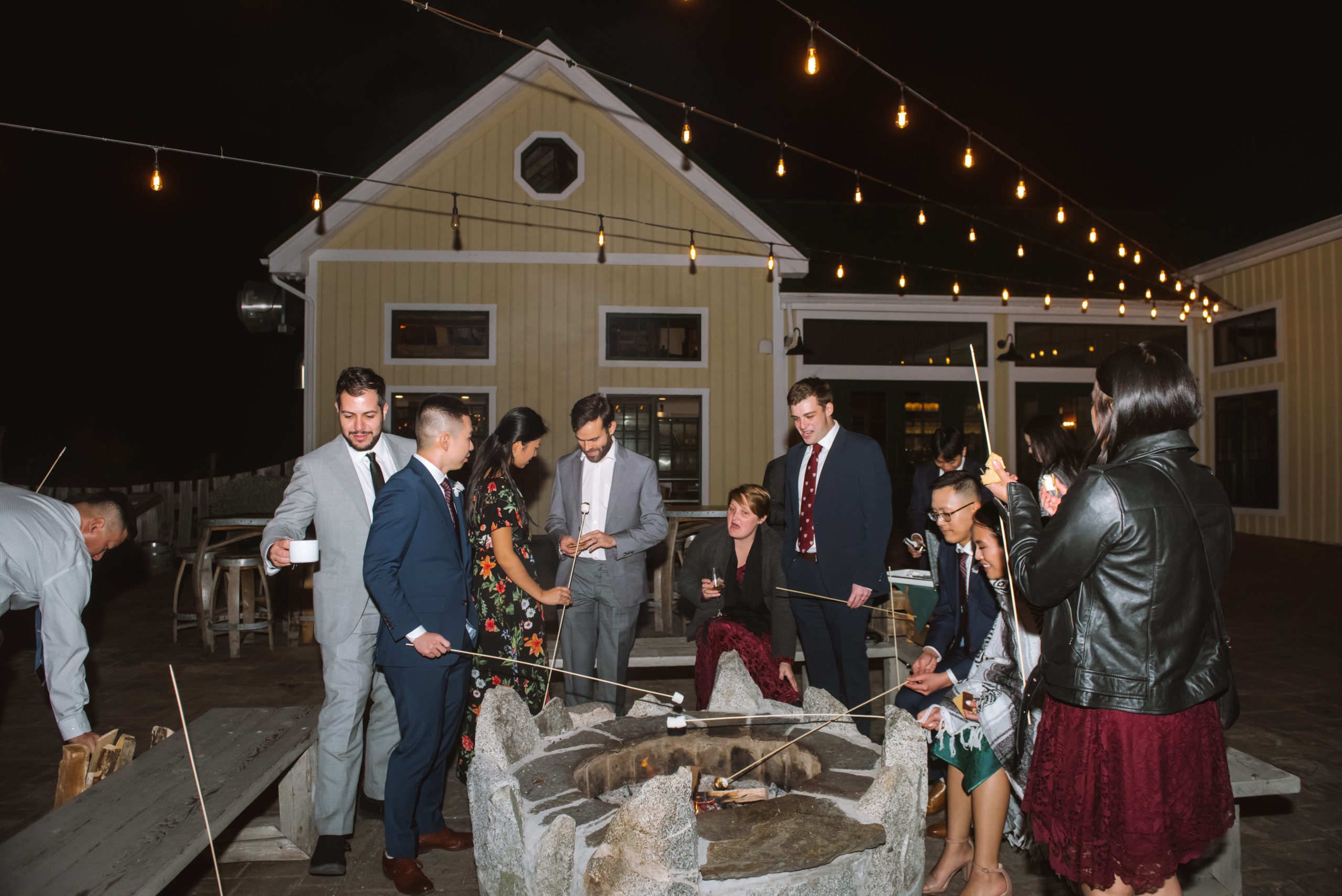 Wedding guests and the two grooms huddled around a bonfire roasting marshmallows for their s'mores. They are all either smiling or in conversation with one another. There are three string light strings hanging above the group.
