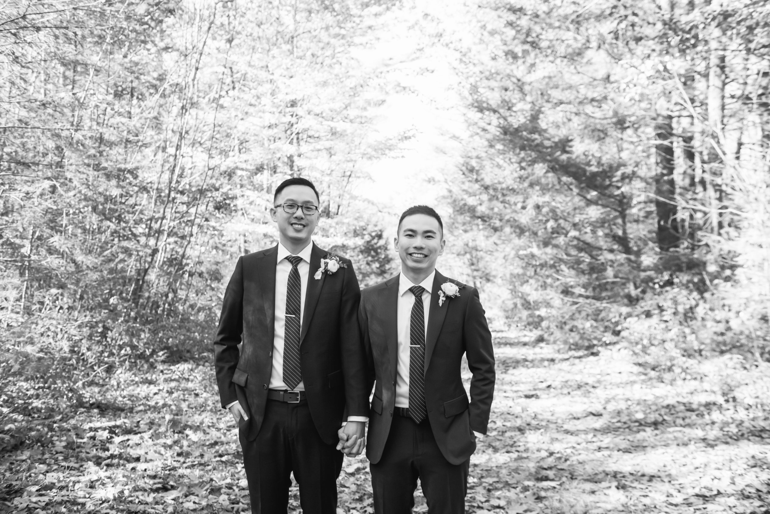 Black and white photo of two grooms standing side by side, holding hands. They are both looking straight to camera and are smiling. They are on a leaf-covered trail and there are trees in the background.