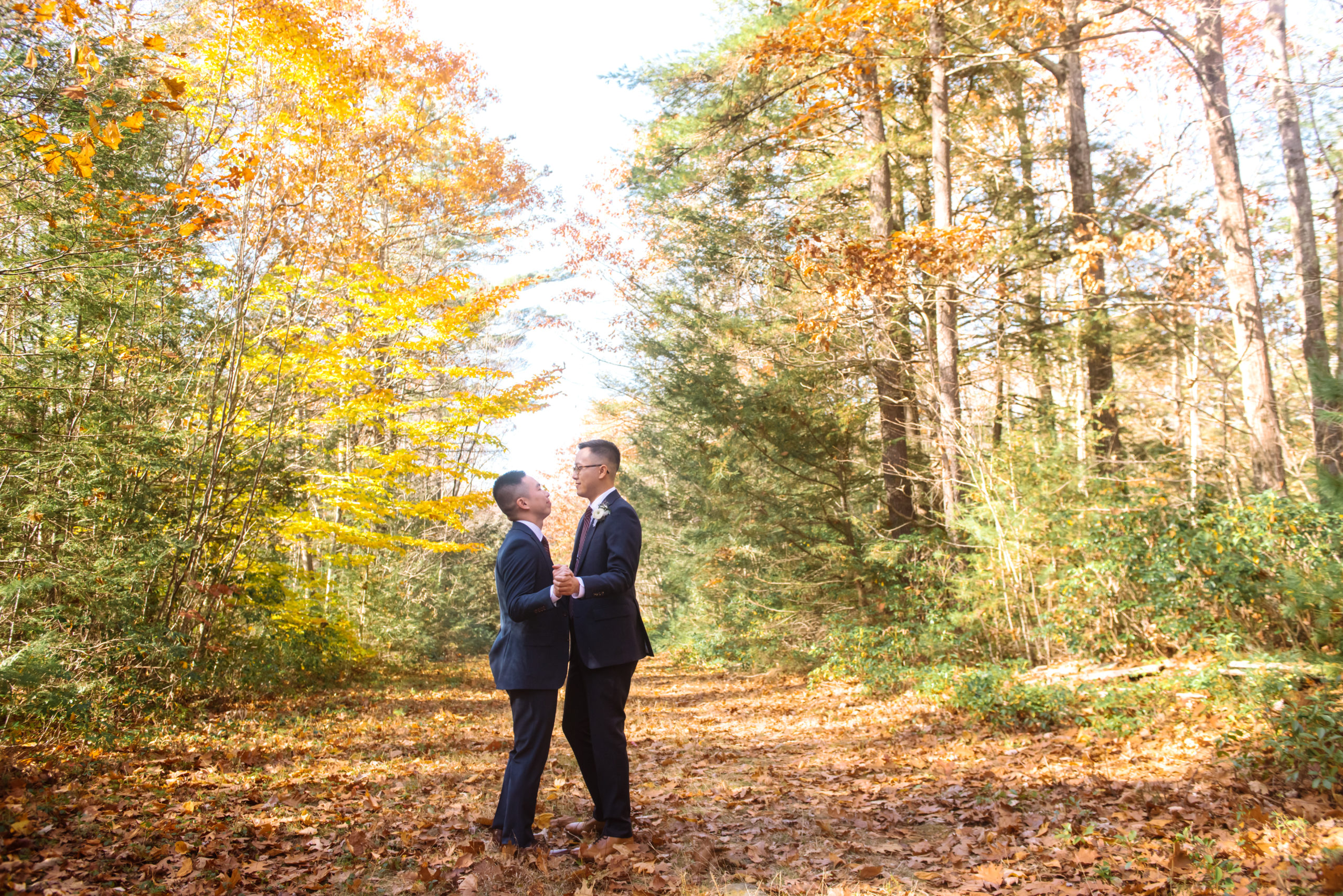 Two grooms slow dancing in each other's arm, facing one another. They are both softly smiling. They are dressed in two different colored dark blue suits, striped burgundy ties, and white boutonnieres. They are in an autumnal forest walking on an autumnal-leaf-covered trail.