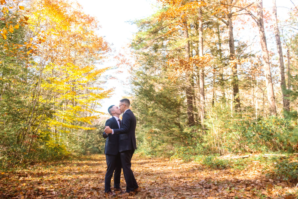 Two grooms kissing, facing one another with their arms wrapped around one another in a slow dance. They are standing in a wide autumn-leaf-covered trail and there are autumnal trees surrounding them.