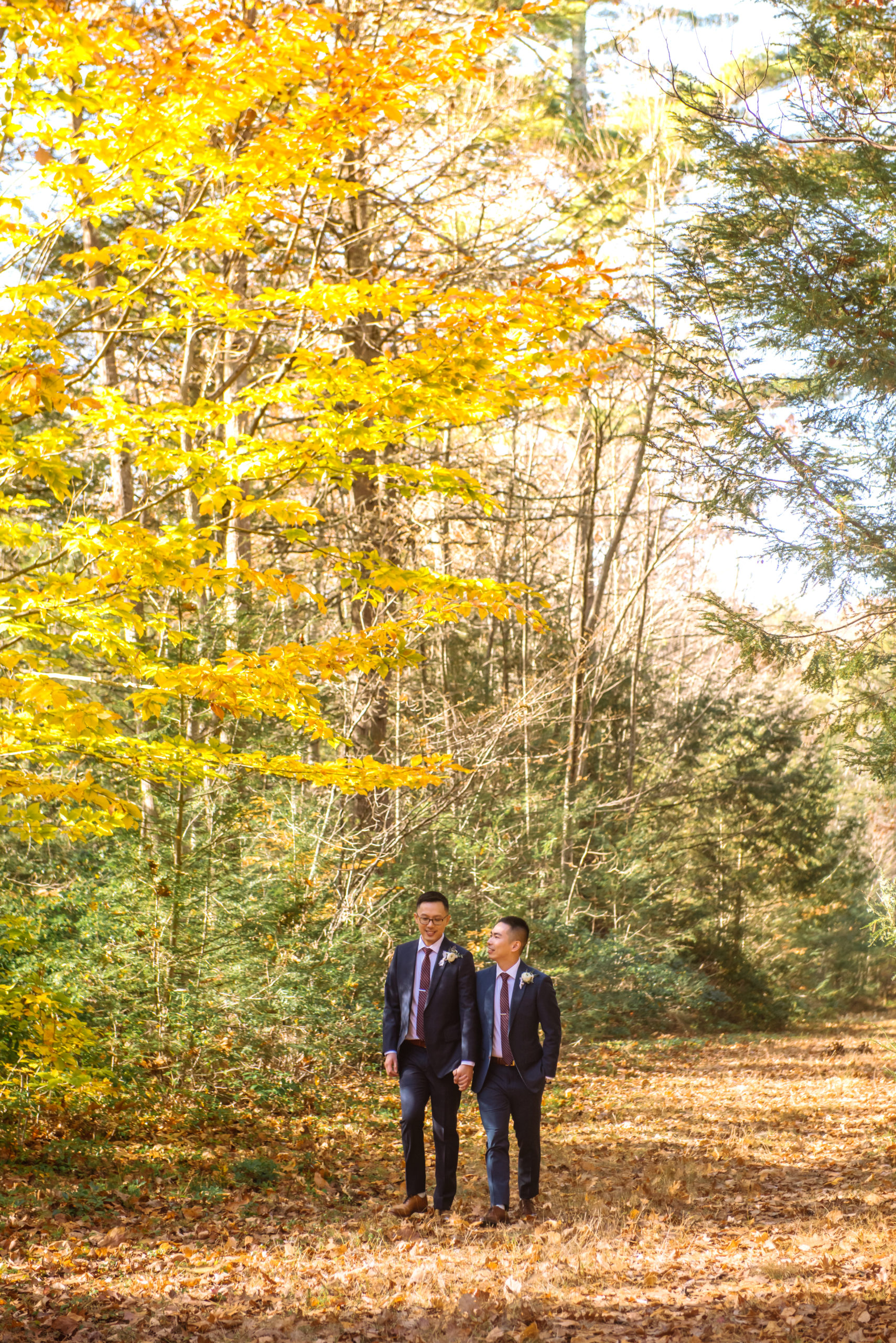 Two grooms walking hand in hand. One is looking down to his left toward the ground and the other is looking up at his soon-to-be husband. They are both smiling. They are dressed in two different colored dark blue suits, striped burgundy ties, and white boutonnieres. They are in an autumnal forest walking on an autumnal-leaf-covered trail.