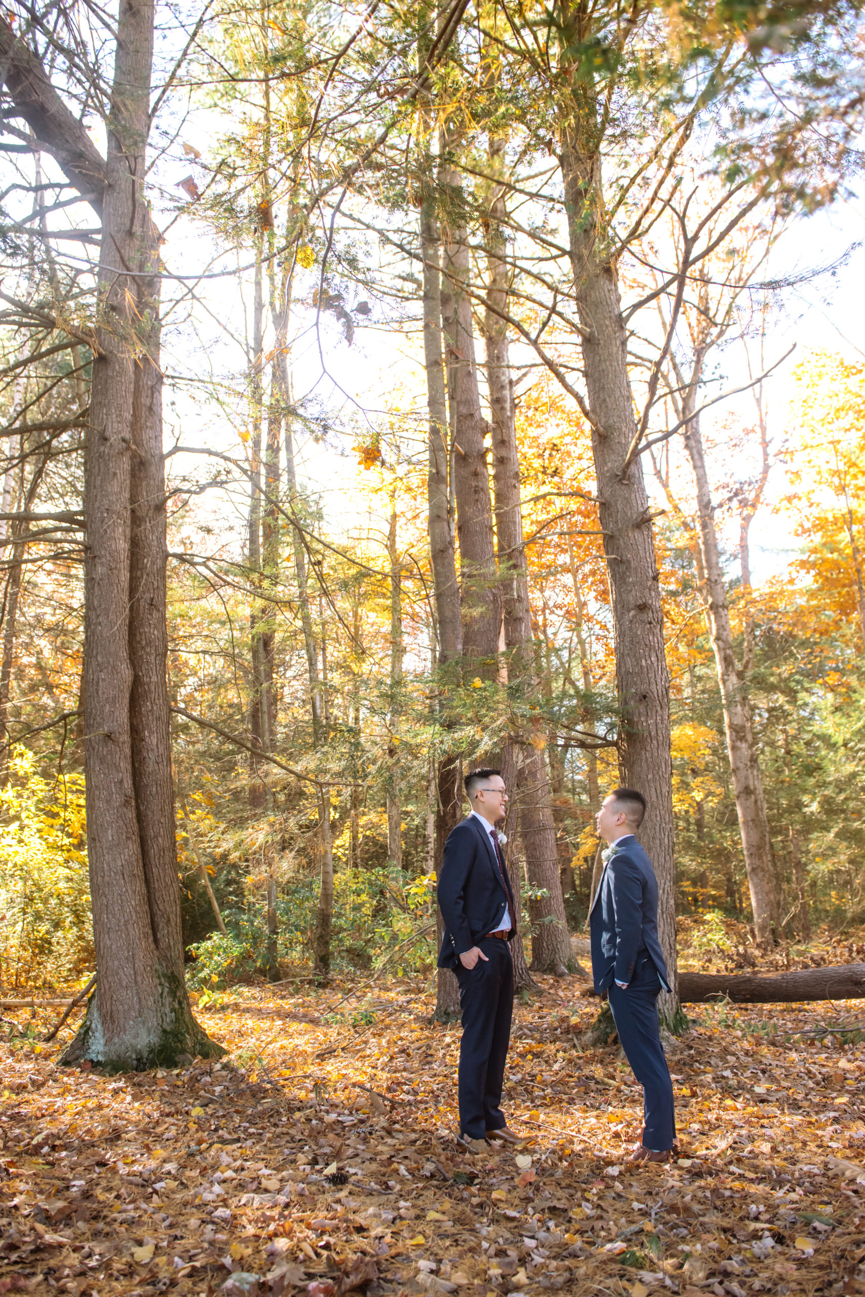 Two grooms standing facing each other with their hands in their pockets. They are both looking at each other, smiling. They are dressed in two different colored dark blue suits. They are in an autumnal forest.