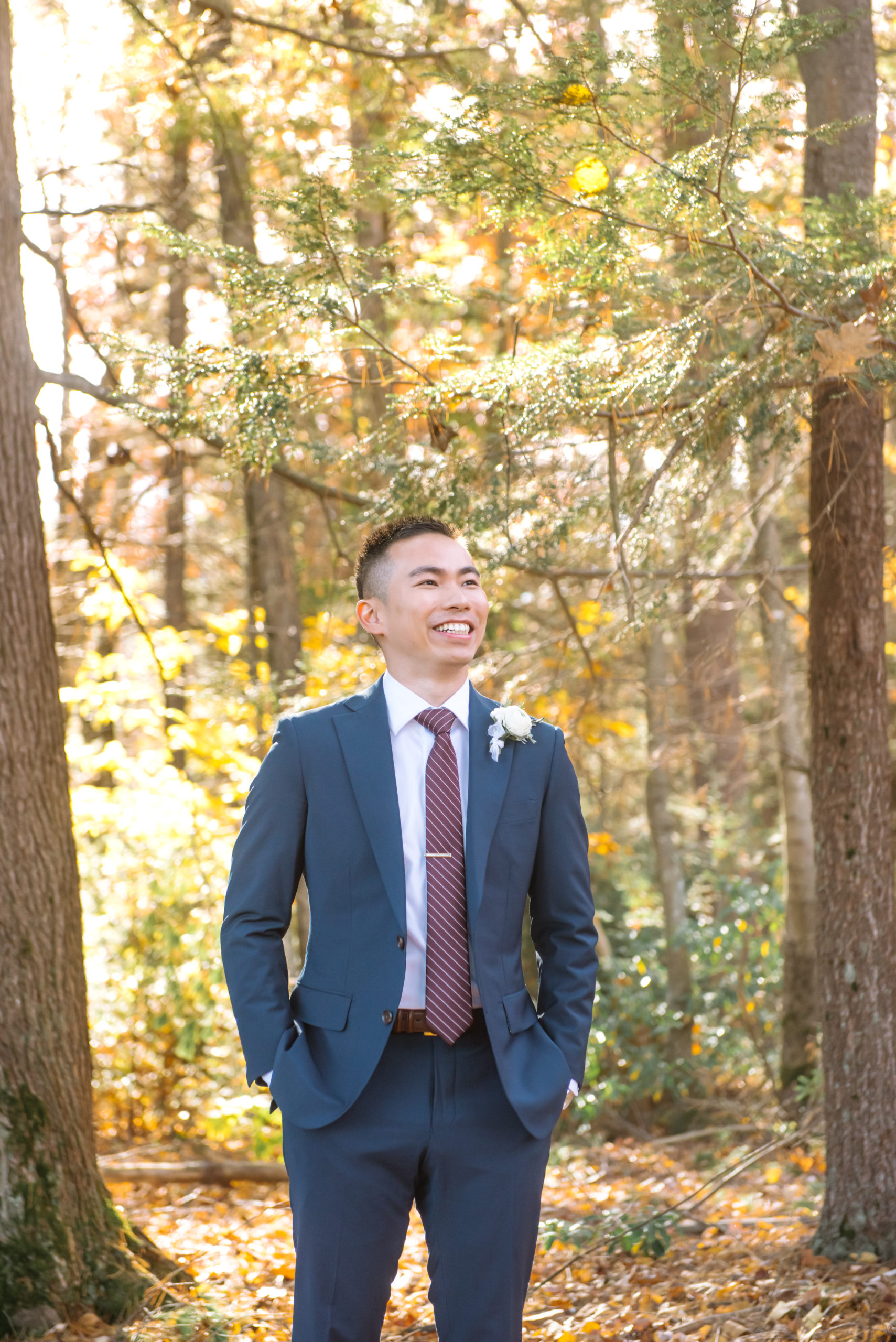 Torso portrait of one of the grooms smiling and looking up and to his left off camera. He is dressed in a dark blue suit with a burgundy striped tie and a white and greenery boutonniere. He is standing in an autumnal forest.