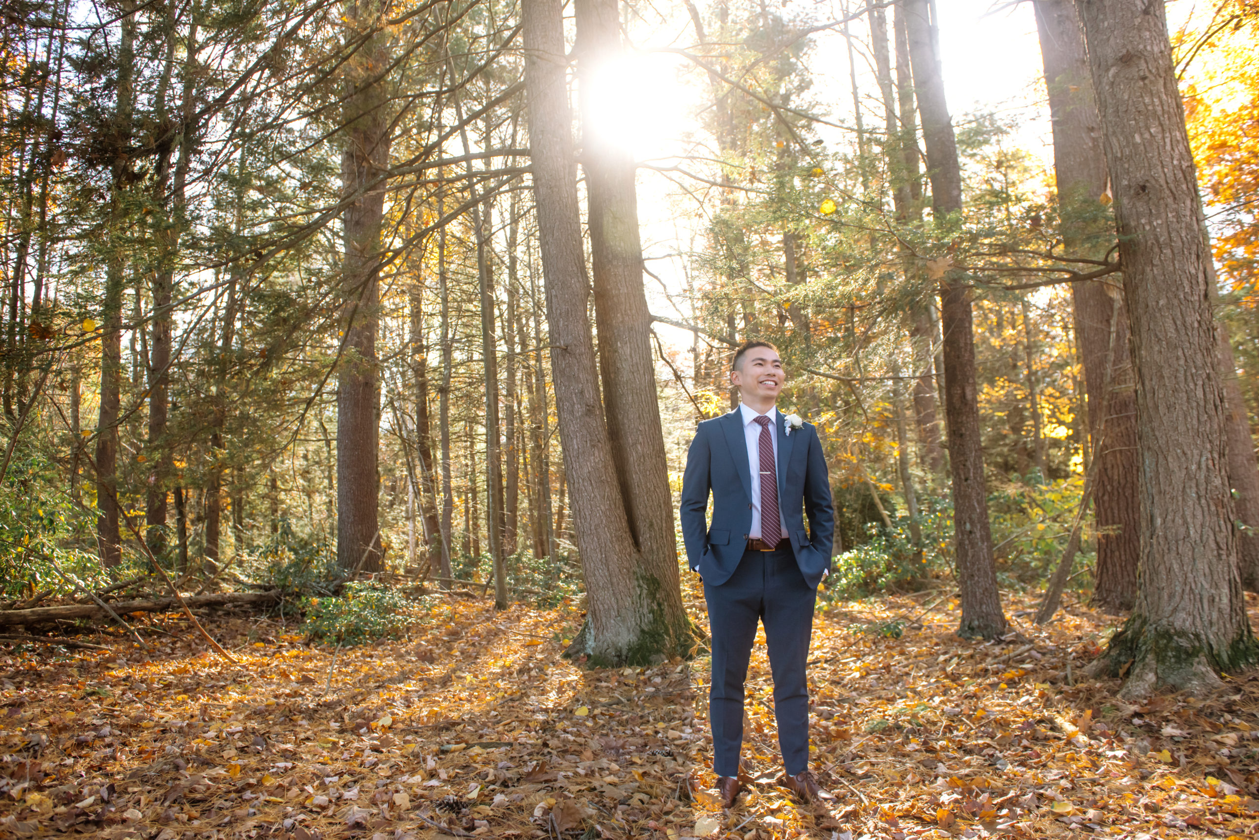 Full body portrait of one of the grooms smiling and looking up and to his left off camera. He is dressed in a dark blue suit, a burgundy striped tie, a white and greenery boutonniere, and brown dress shoes. He is standing in an autumnal forest with the sun shining through the trees.