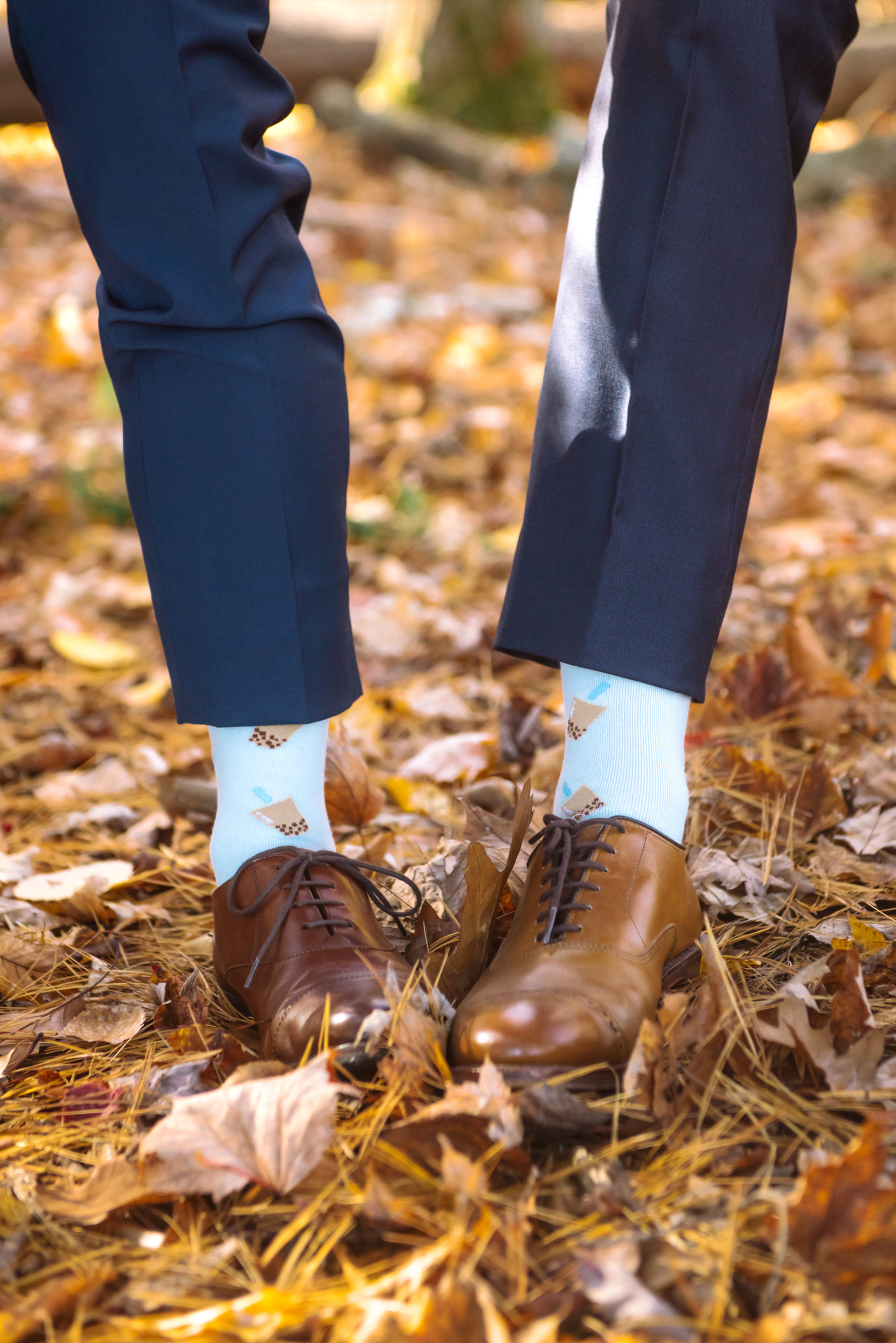 Detail of the groom's wedding shoes and milk tea bubble tea socks. In view is one of each groom's leg. They are standing atop autumnal leaves.
