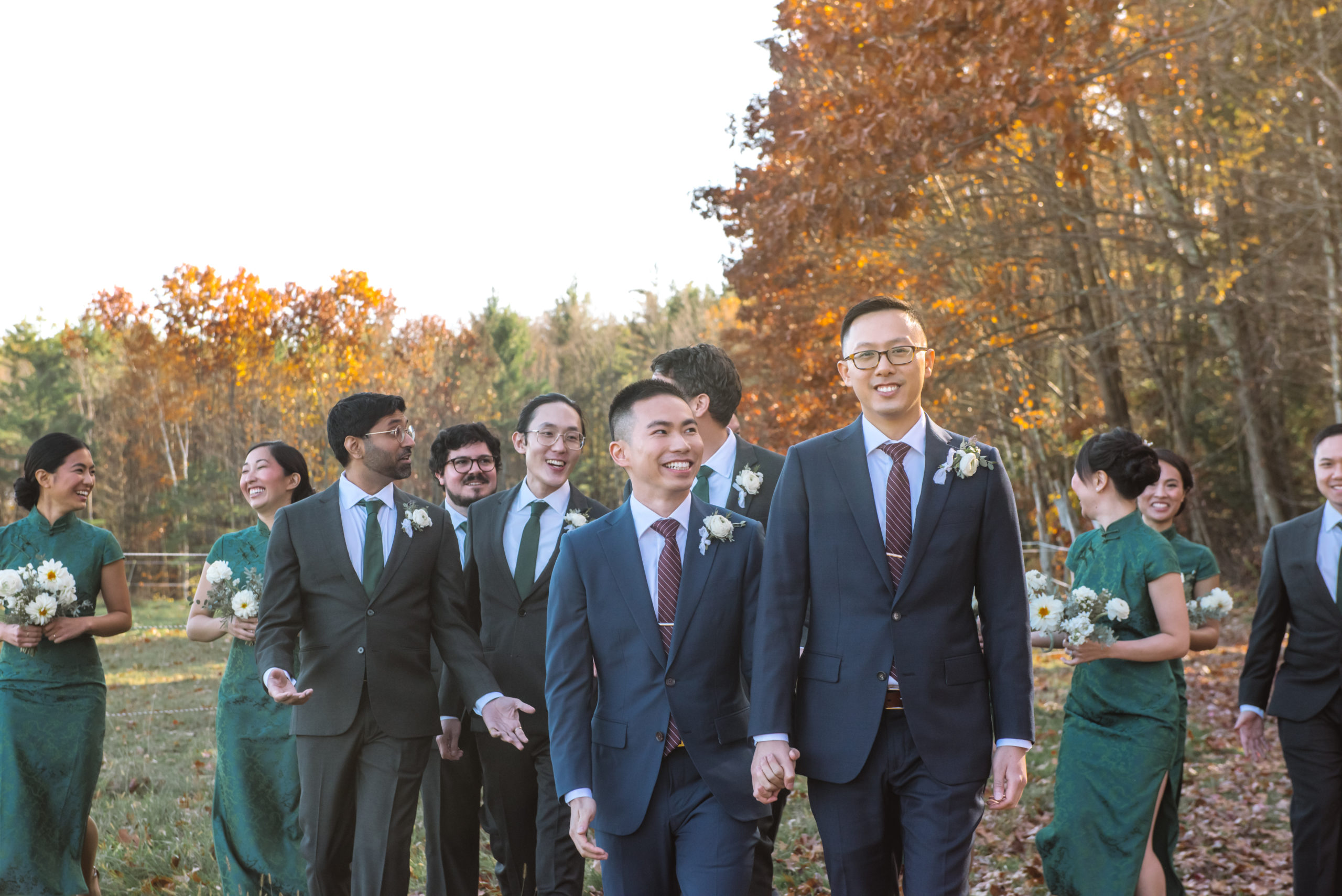 Two grooms walking hand in hand, one looking directly to camera and the other looking up to his partner. Their wedding party is following them behind and everyone is smiling. Autumnal trees are in the background.