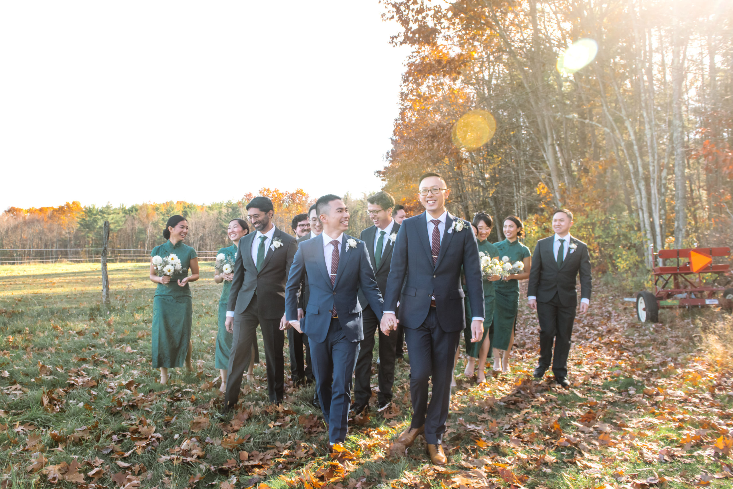 Full body portrait of two grooms walking hand in hand, one looking directly to camera and the other looking up to his partner. Their wedding party is following them behind and everyone is smiling. The women are dressed in dark green qi paos and are holding white bouquets. The men are dressed in black suits with matching dark green ties and white boutonnieres. Autumnal trees are in the background with the sun shining through them.