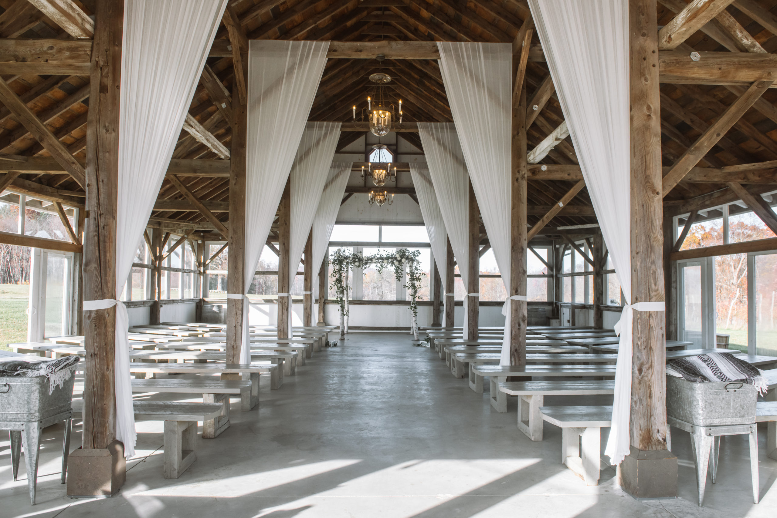 Ceremony location featuring barn beams covered in decorative white sheer curtains. There are aisles of empty wooden benches for the guests to sit on. It is a light-filled area full of glass windows and glass doors. At the end of the aisle is the alter which is comprised of birch tree branches and decorated with greenery and white florals.