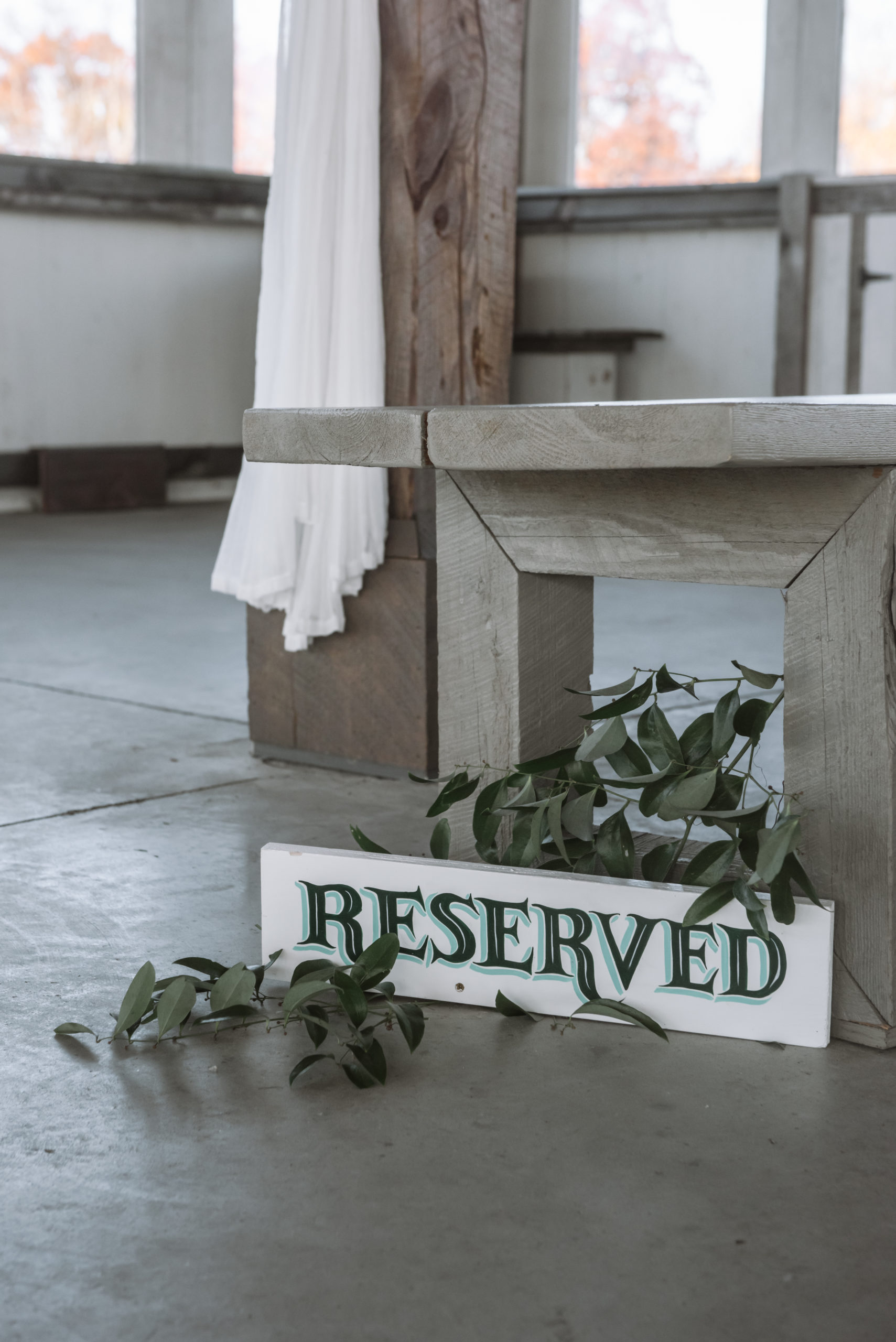 Detail of a ceremony sign labeled "RESERVED" which is set on the ground among some greenery by one of the wooden aisle benches.