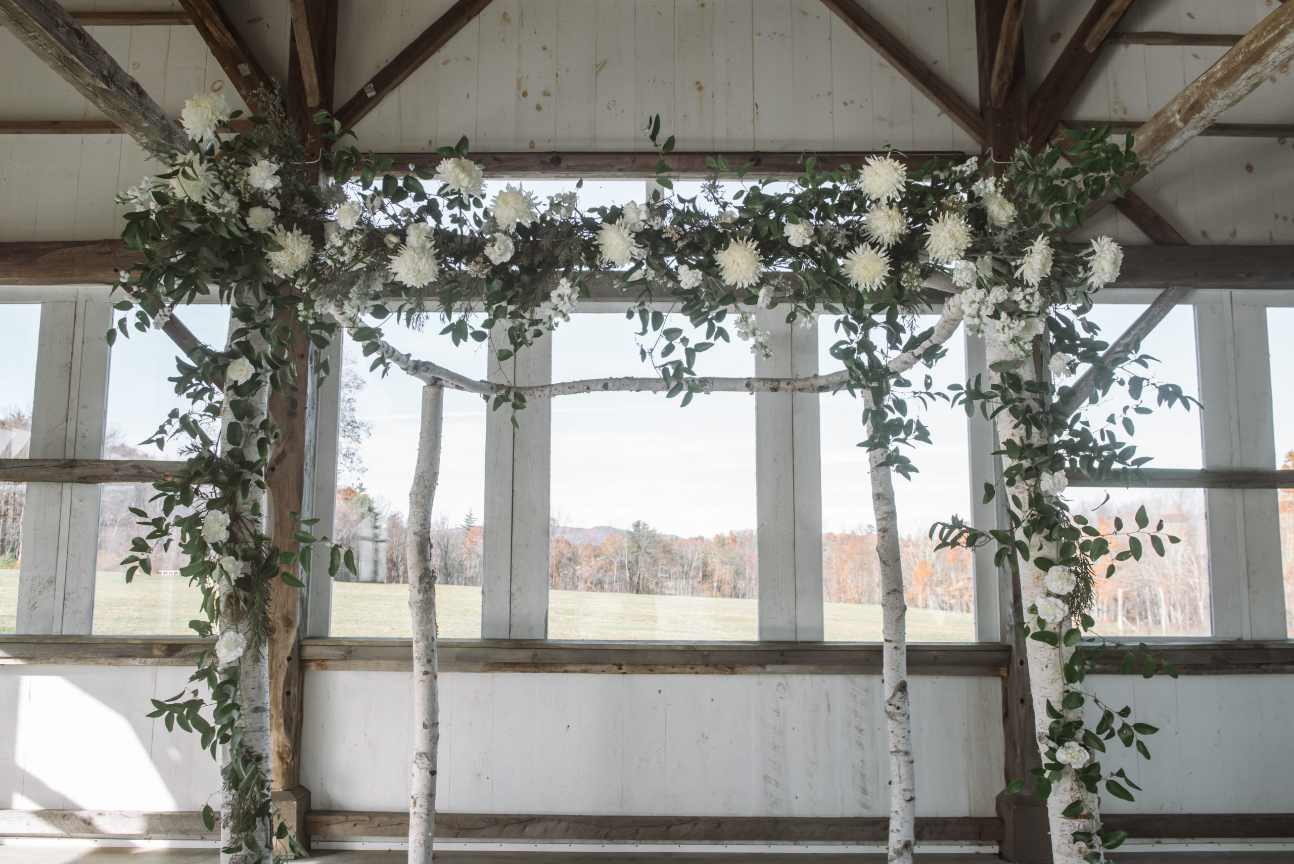 An alter in a ceremony location that is decorated with birch trees, greenery, and white florals. There are wooden beams in the location. There are windows behind showing an autumnal forest in the background.