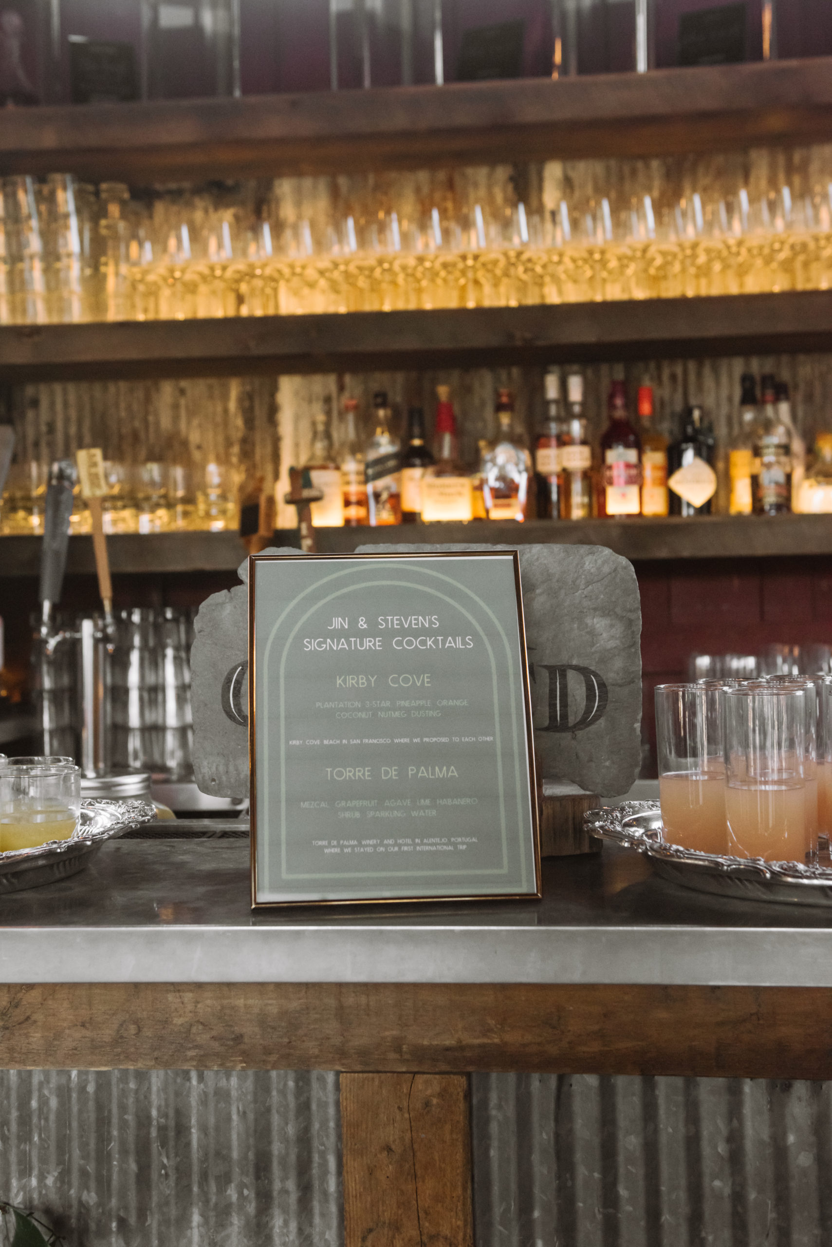Detail of the groom's signature cocktail menu. It is set atop the bar counter and flanked by said signature cocktails. There are multiple wine glasses and various bottles of liquor on the shelves in the background. The two signature cocktails are called "Kirby Cove" and "Torre de Palma".