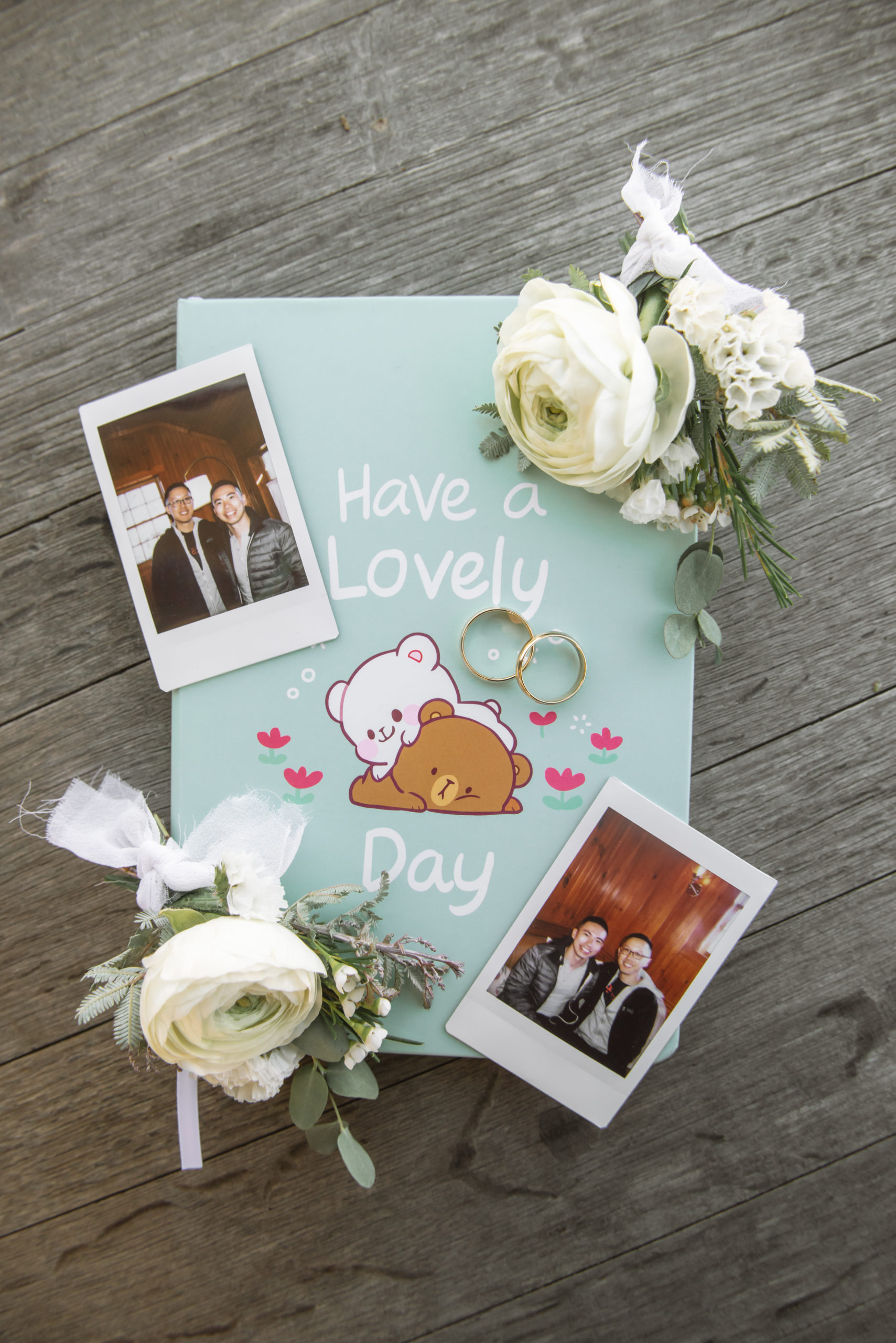 Detail of the grooms' two gold wedding bands, two boutonnieres, two polaroid photos of the grooms, and the wedding vow book which has the words "Have a Lovely Day" written on it. The journal has illustrations of one white bear and one brown bear and they are flanked by pink flowers and white sparkle/bubble motifs.