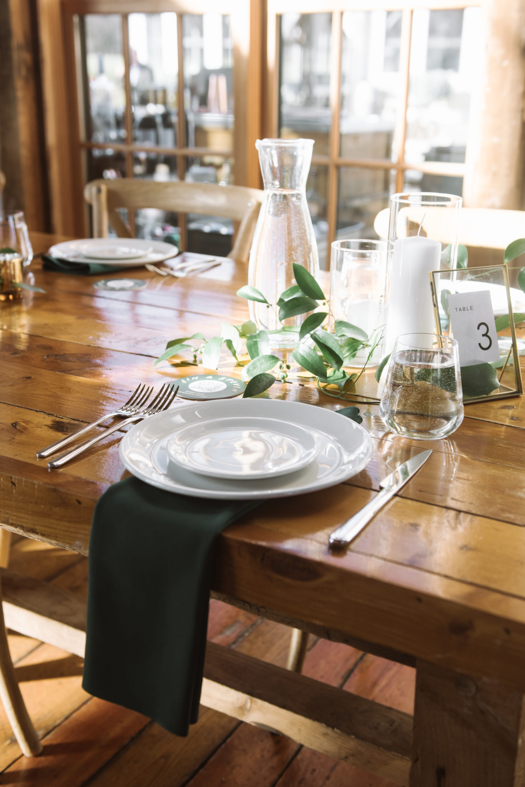 An individual place setting comprised of two white plates on top of one another flanked by two forks and one knife. There is a dark green napkin below the plates. There is greenery as part of the centerpiece and the table is made of wood. The sun is shining brightly in the background.