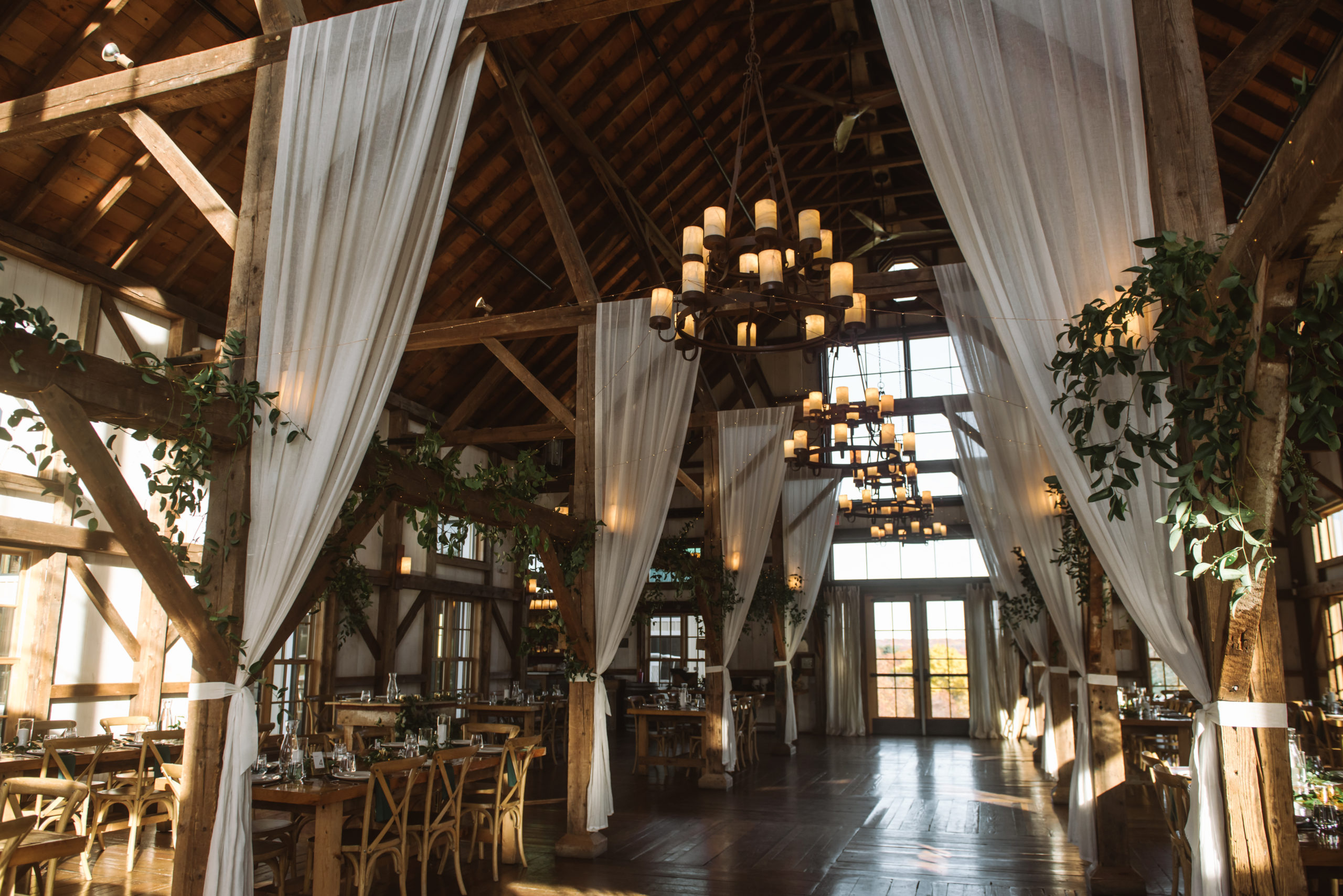 Renovated barn reception venue featuring multiple wooden tables and chairs. There are hanging wide candle-like chandeliers that are lit. Sheer white curtains decorate the wooden beams alongside greenery.