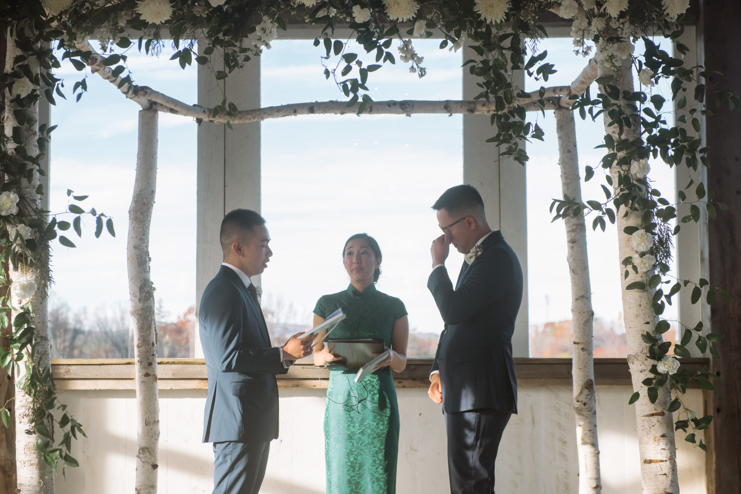 Two grooms mid-ceremony. One is reading his vows and the other is wiping away tears. The officiant is looking towards the groom who is reciting his vows. She is dressed in a green qi pao and the grooms are dressed in two different shades of dark blue suits. The alter is decorated with birch trees, greenery, and white florals.