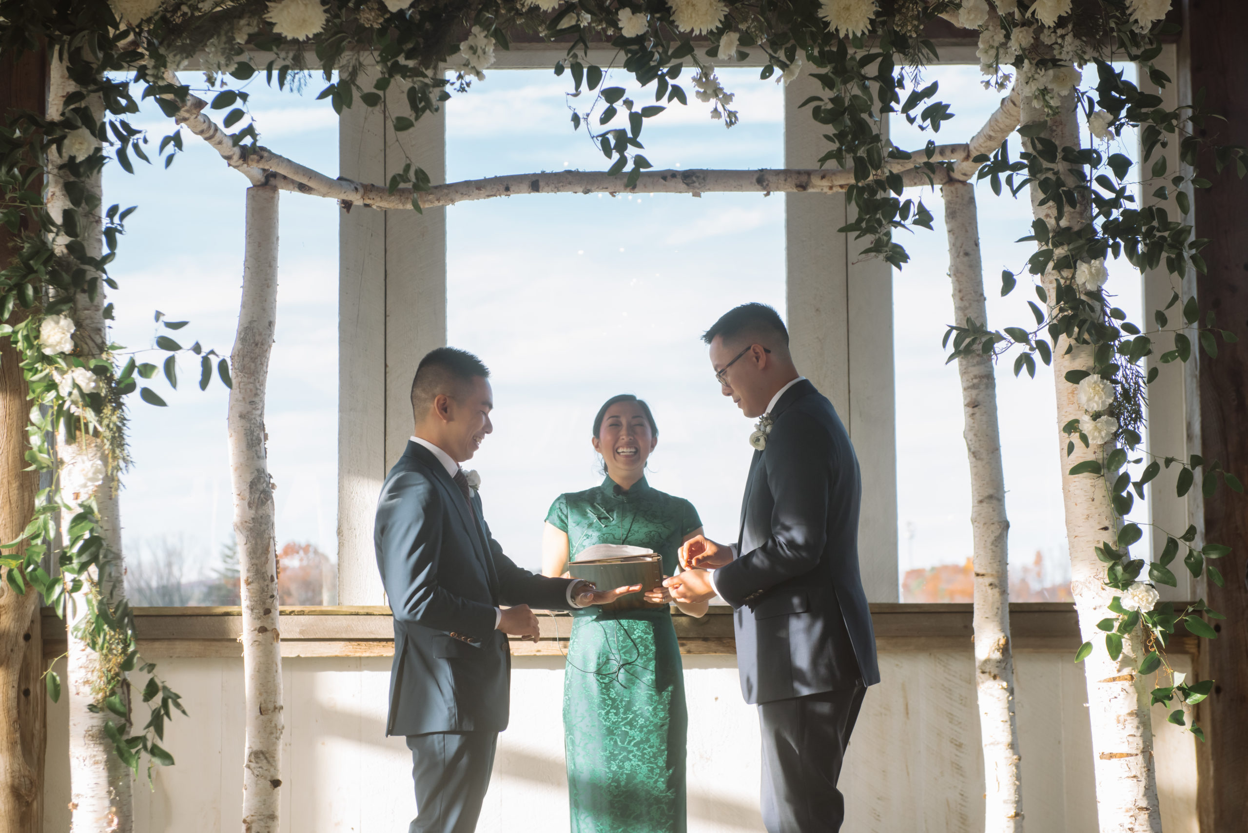 Two grooms mid-ceremony. One is about to place the wedding band on the other's hand. The officiant is smiling wide. She is dressed in a green qi pao and the grooms are dressed in two different shades of dark blue suits. The alter is decorated with birch trees, greenery, and white florals.