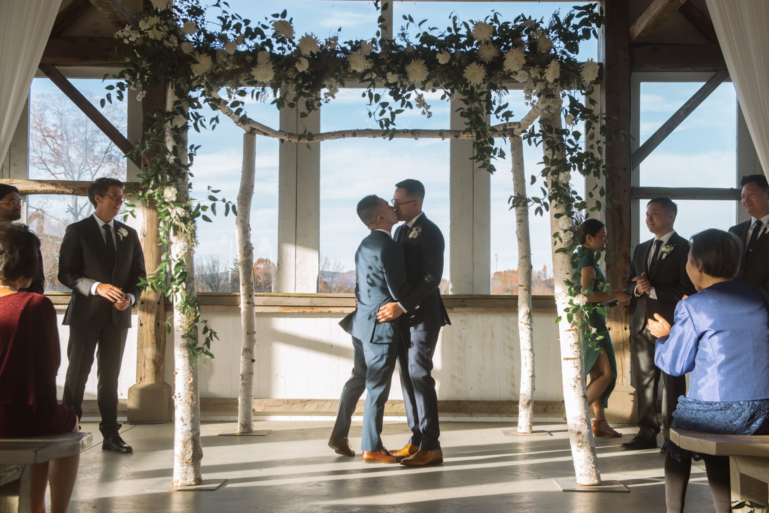 Two grooms kissing at the end of the ceremony. The grooms are dressed in two different shades of dark blue suits. They are surrounded by their wedding party and visible guests are one of the groom's mother and the other groom's grandmother who is clapping. The alter is decorated with birch trees, greenery, and white florals.