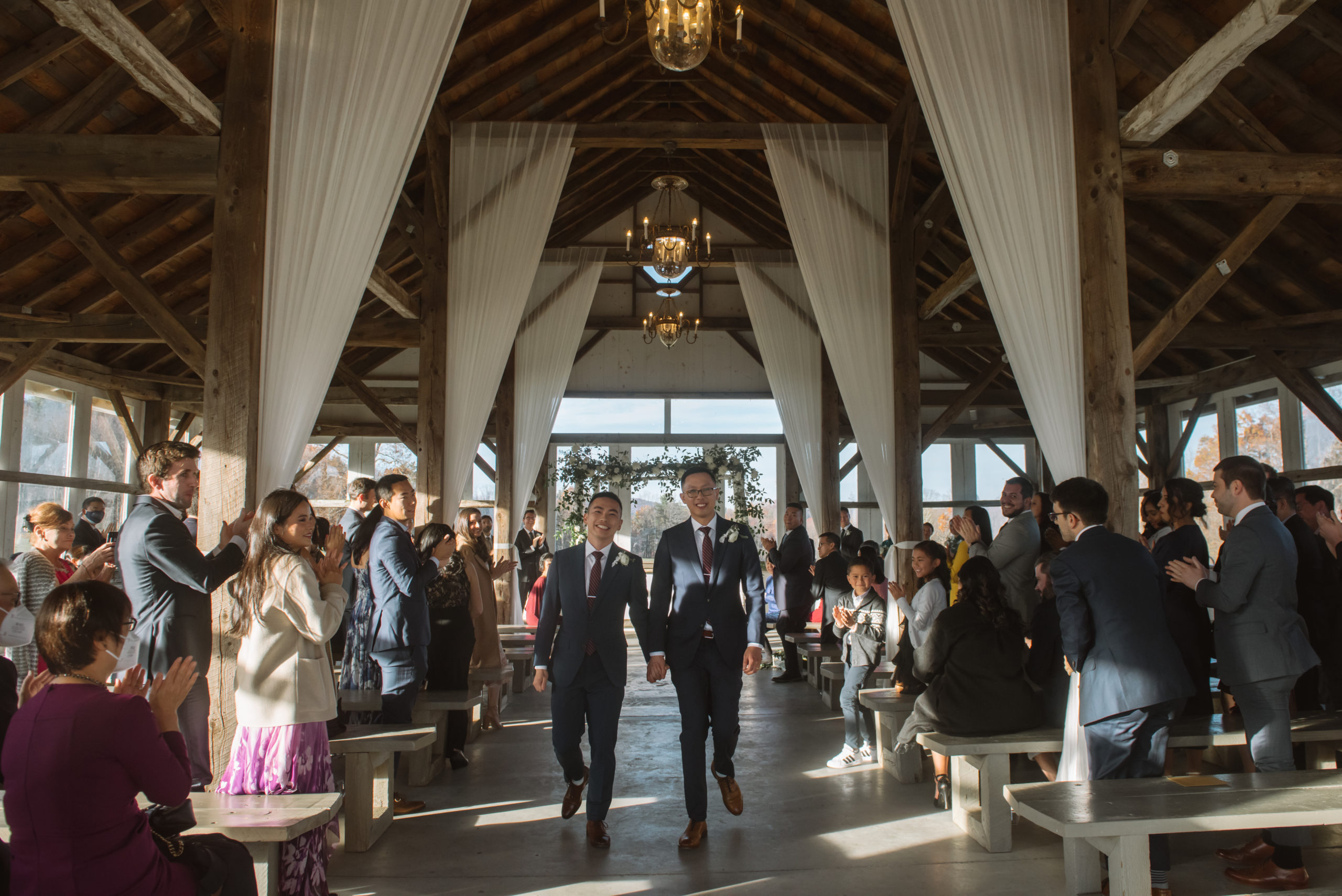 Two grooms walking hand in hand with big smiles right after their ceremony. All of their guests are smiling and clapping for the happy couple. The ceremony location is a barn-style indoor area with big wooden beams and wooden benches for the guests. The beams are decorated with white sheer curtains.