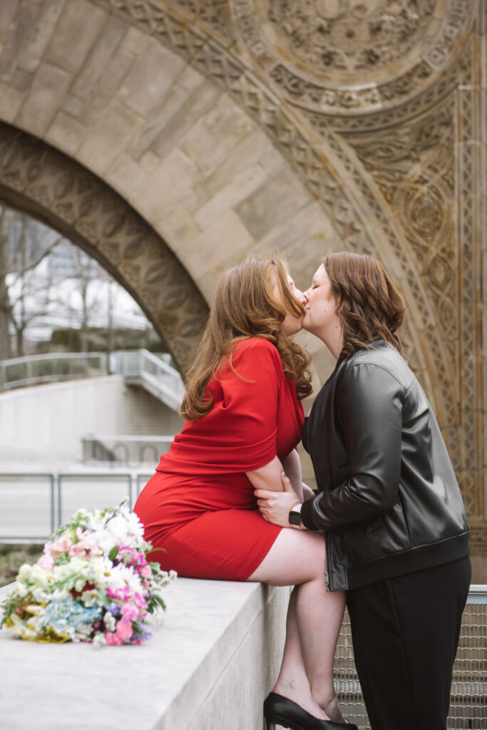 Two brides kissing, facing one another with Rya's hands on Britt's arms. Rya is standing and Britt is sitting atop a stone bench. They are in front of an intricately carved stone archway in Chicago.