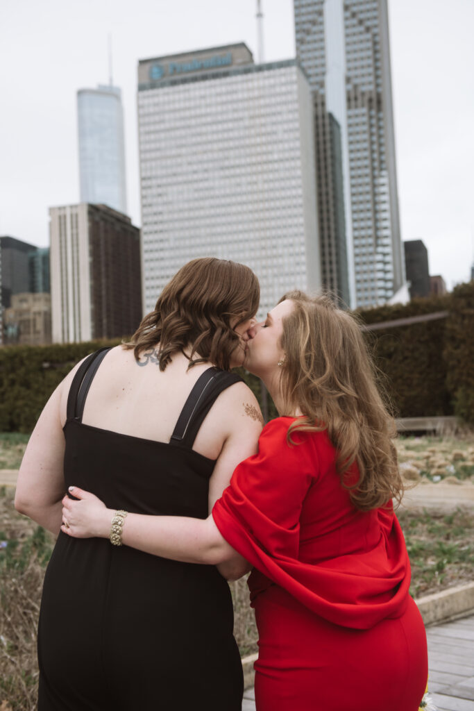 Two brides kissing, one with her arm wrapped around the other. Their backs are to the camera and they are standing in a garden with Chicago skyscrapers in the background.