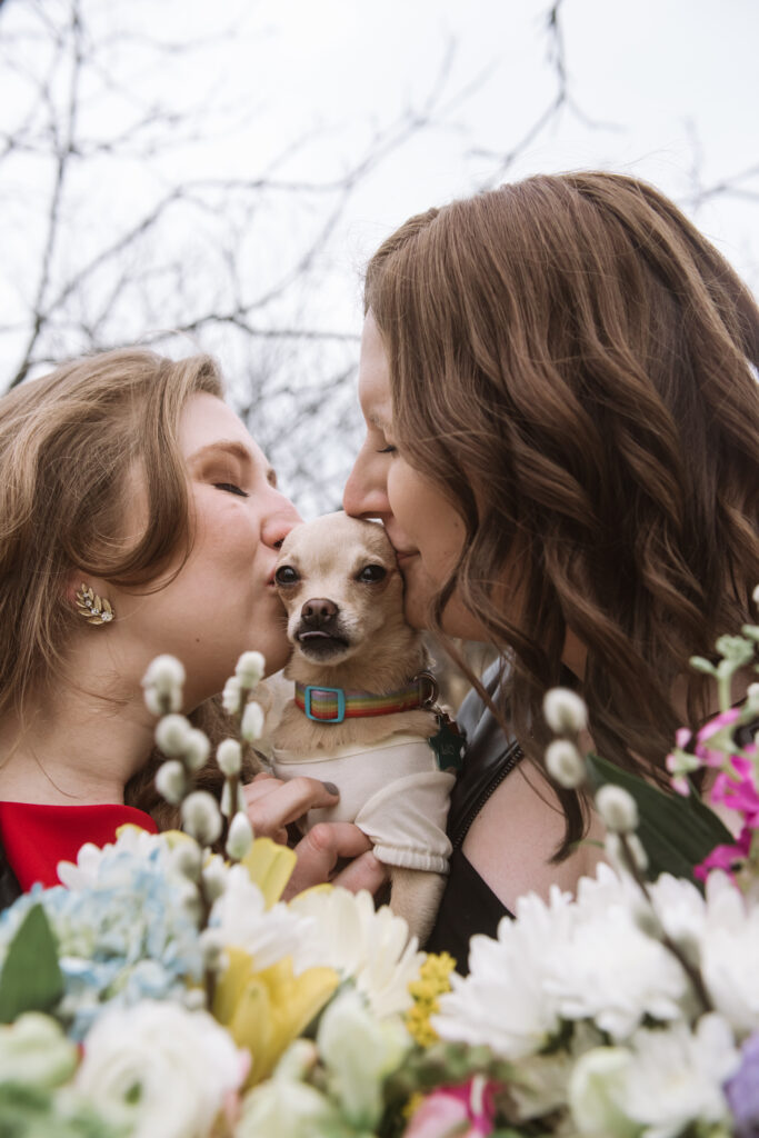 Close up of two brides holding their little Chihuahua in between them. Their Chihuahua is facing the camera directly. She is dressed in a shirt with a rainbow colored collar. Her eyes are wide open and her tongue is barely visible through her barely open mouth. Both brides are giving their dog a kiss, eyes closed, from either side of the dog. Their bouquets are visible in the bottom foreground with trees/tree branches in the background.