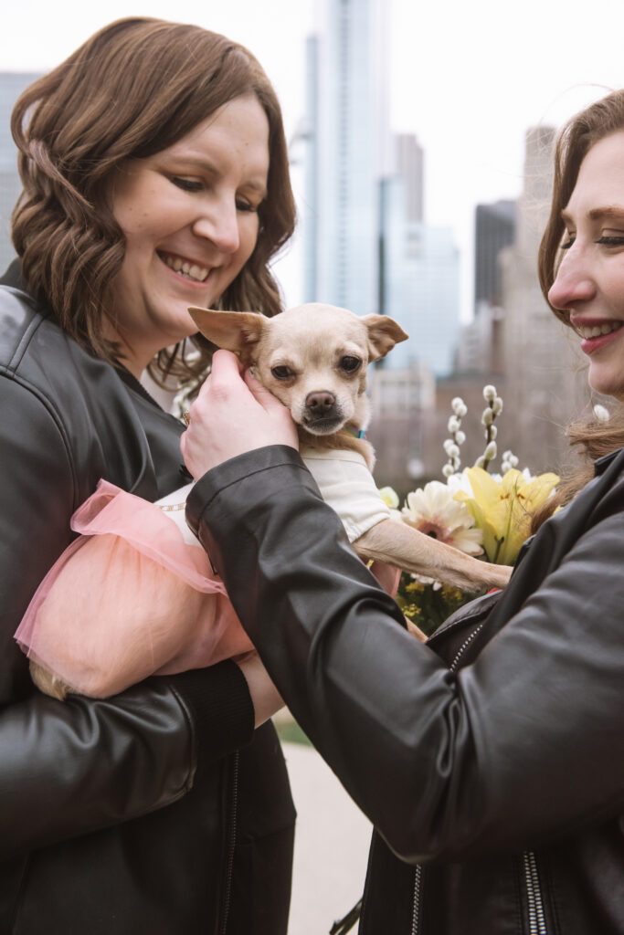 Close up of two brides holding their little Chihuahua in between them. Their Chihuahua is facing the camera directly. She is dressed in a shirt + tutu outfit. One lower right canine is visible through her closed mouth while she is looking off camera. Both brides are looking at their dog with open-mouthed smiles.