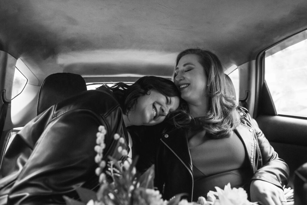 Black and white photo of two brides sitting in the back of a cab, one has her head on the other's shoulder. They are both smiling with their eyes closed. Their bouquets are slightly visible from resting on their laps.